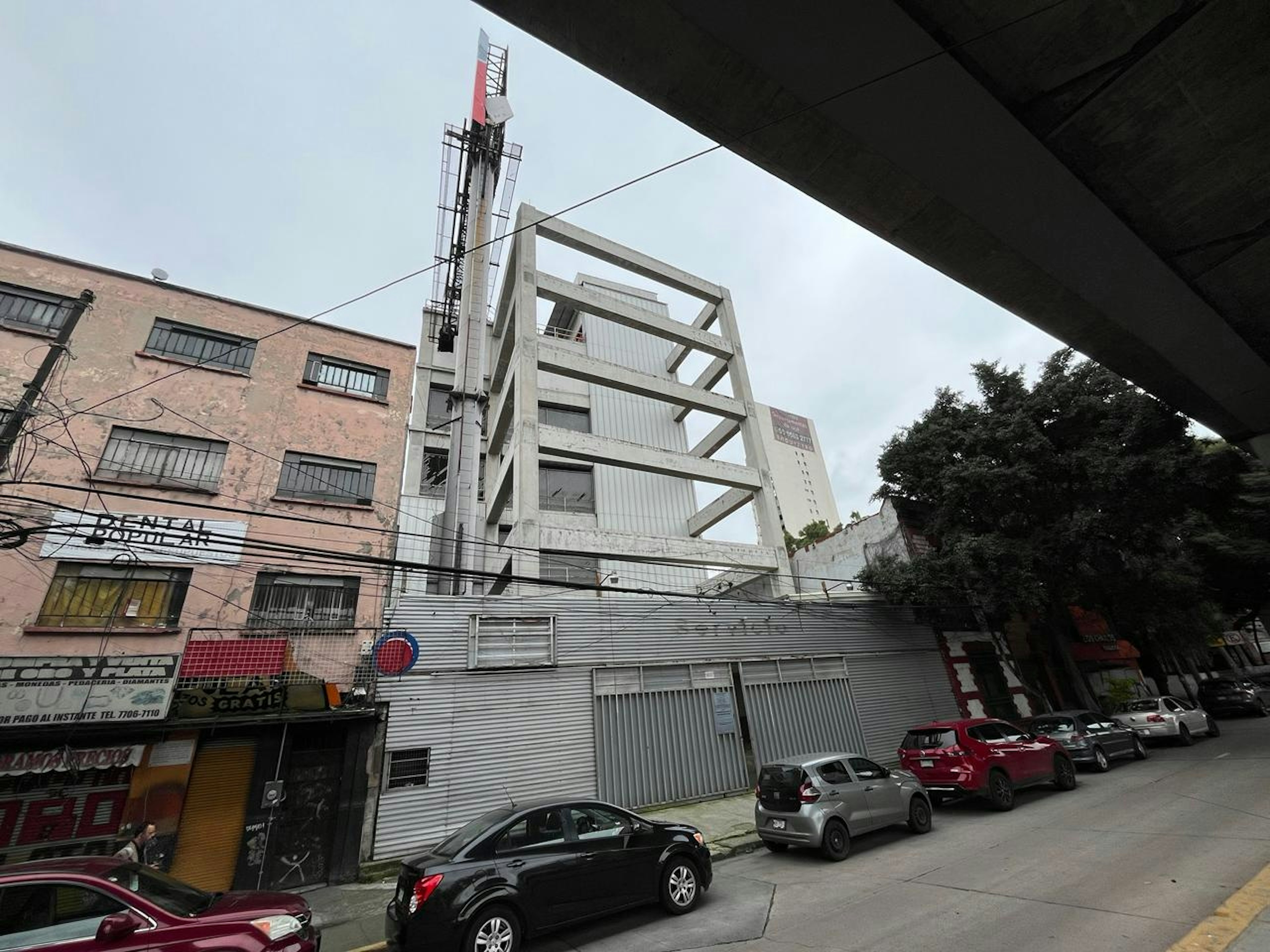 La imagen muestra un área urbana con edificios de diferentes estilos arquitectónicos. Se destaca una estructura moderna de concreto con un diseño esquelético, junto a un edificio más antiguo de ladrillo rosa. En primer plano hay una calle con varios automóviles estacionados. Se observa también una torre de telecomunicaciones. El entorno sugiere una zona en desarrollo o renovación urbana, mezclando lo nuevo con lo antiguo.