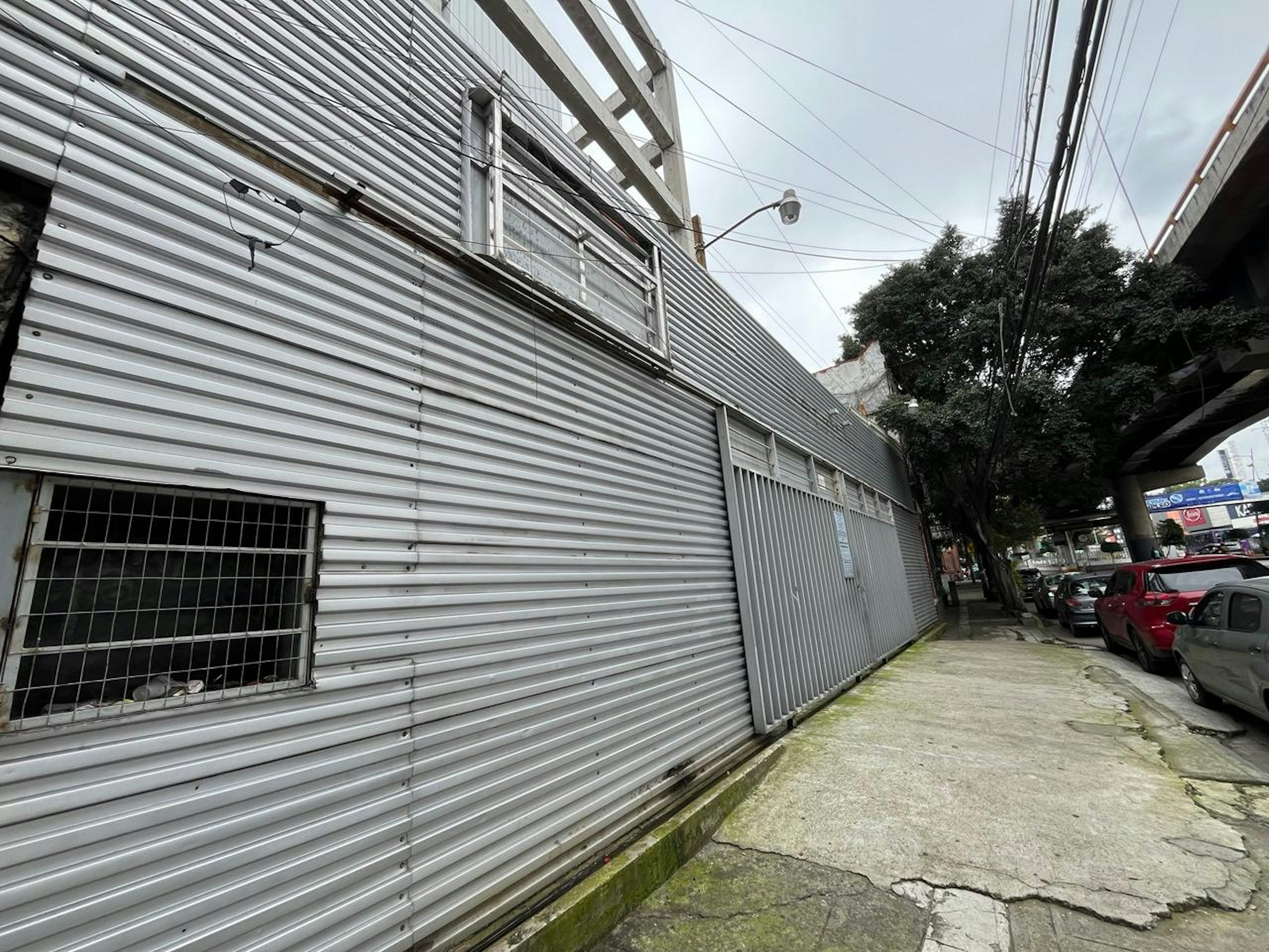 La imagen muestra un edificio industrial con fachada de láminas metálicas corrugadas, típico de bodegas o naves industriales. Se observa una estructura de dos pisos con ventanas enrejadas y un sistema de cámaras de seguridad. El inmueble está ubicado en una zona urbana, con árboles y vehículos estacionados en la calle. La acera muestra signos de desgaste, indicando un área posiblemente comercial o industrial.