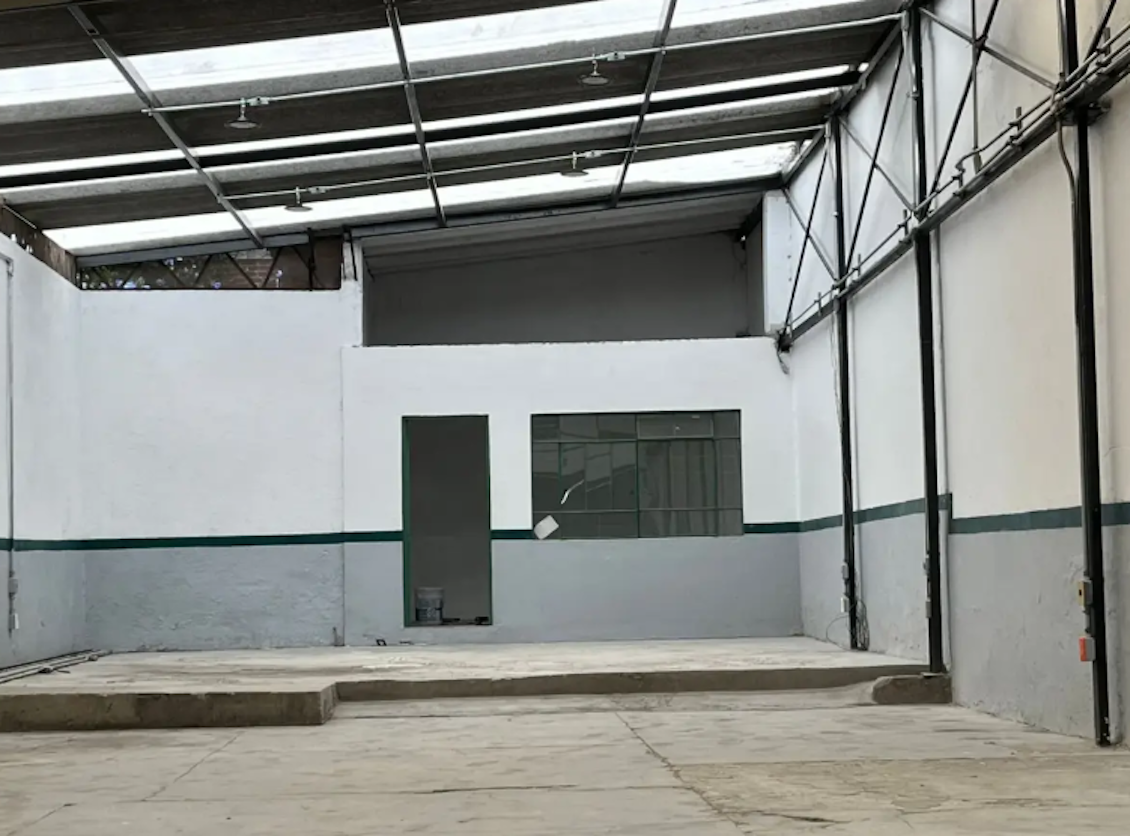 Esta imagen muestra un espacio industrial o de almacenamiento en construcción o renovación. Se observa un techo alto con estructura metálica y tragaluces, paredes blancas con una franja verde, un piso de concreto y una puerta y ventana al fondo. El espacio es amplio y parece estar en proceso de acondicionamiento, ideal para uso comercial o industrial.
