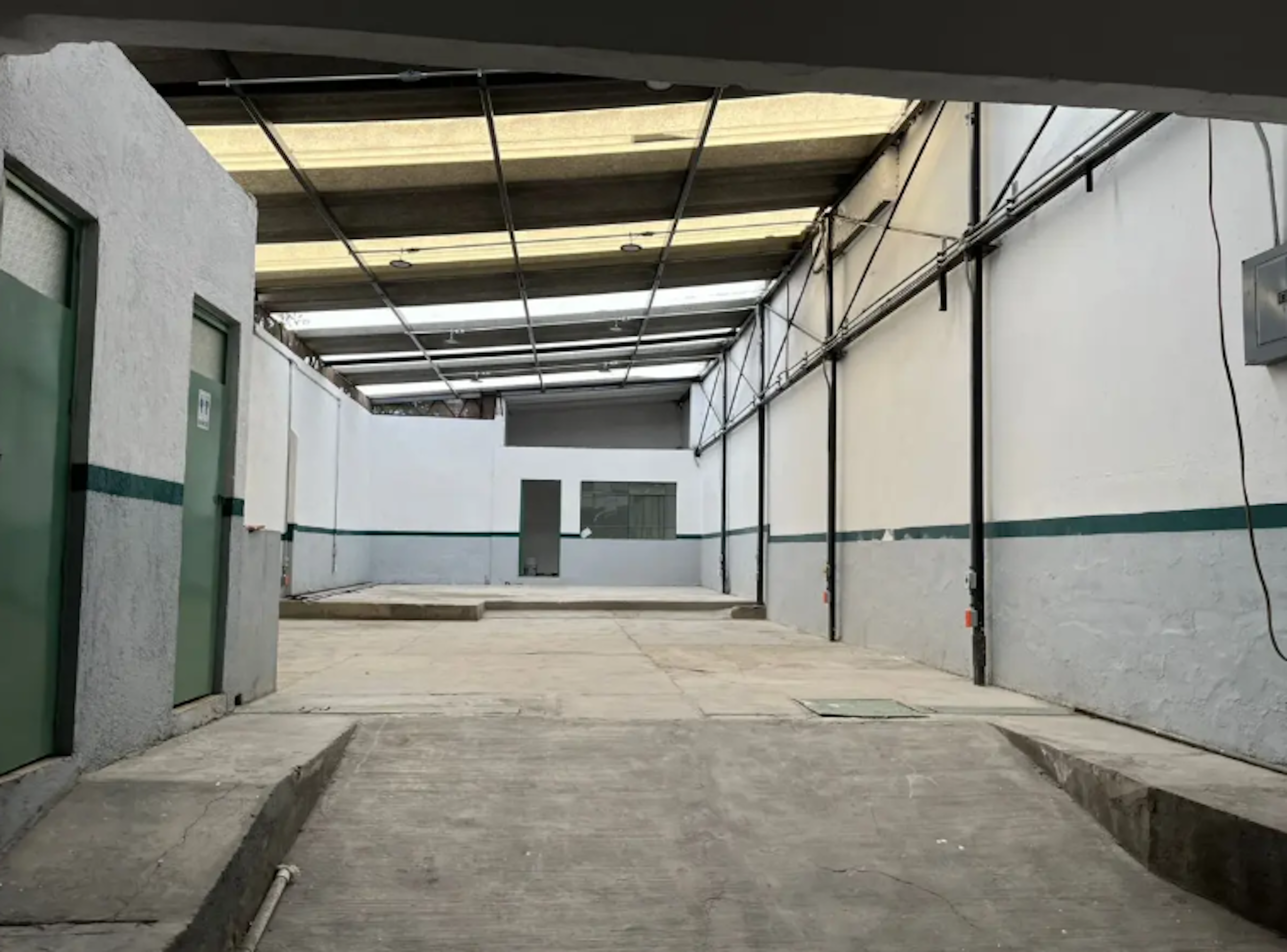 La imagen muestra un espacio interior industrial o comercial en construcción o renovación. Tiene paredes blancas con una franja verde, piso de concreto y un techo de estructura metálica con claraboyas. Se observan puertas verdes y una área amplia que podría ser utilizada como almacén, taller o espacio de trabajo. El lugar parece estar en proceso de acondicionamiento, lo que sugiere potencial para diversos usos comerciales o industriales.