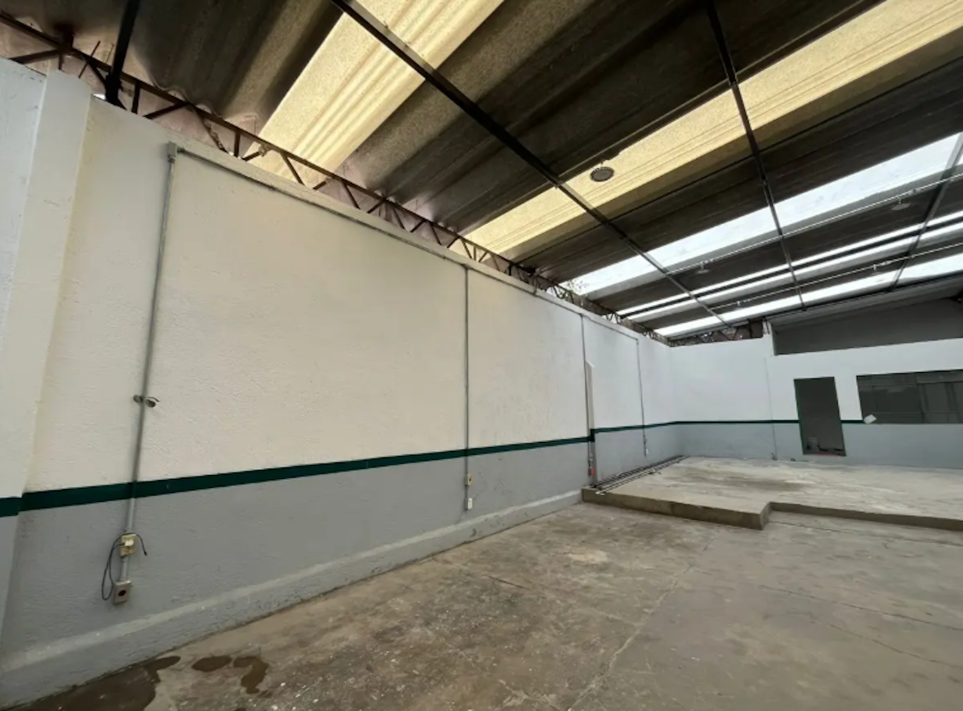 La imagen muestra un espacio industrial o comercial vacío con techo de lámina, paredes blancas y un zócalo verde. El piso es de concreto sin acabados. Se observan algunas instalaciones eléctricas básicas y una puerta al fondo. El área parece estar en proceso de remodelación o preparación para su uso, ideal para almacenamiento o como área de trabajo.