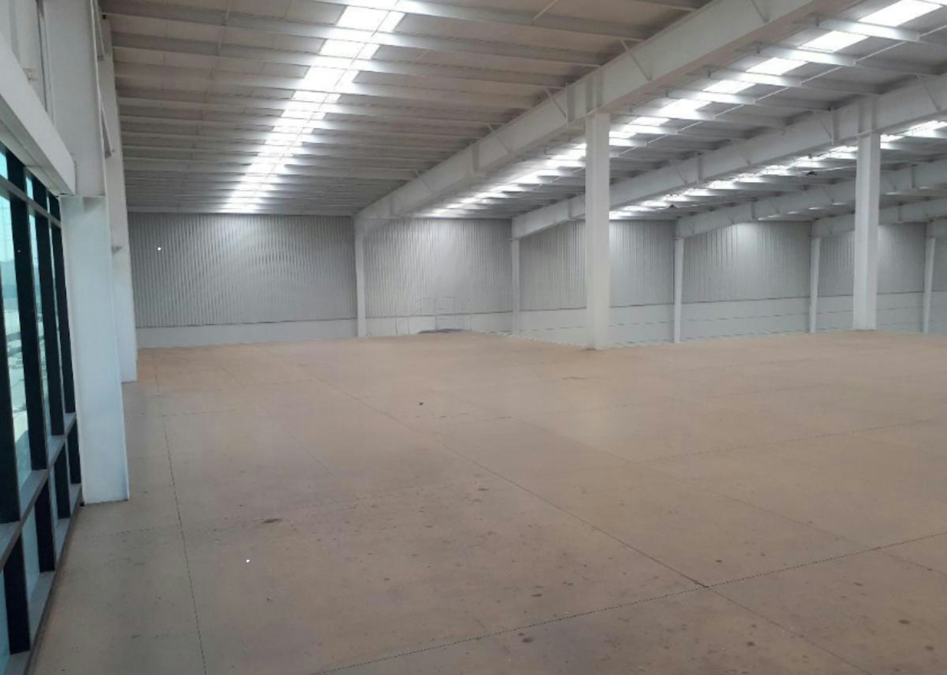 La imagen muestra un amplio espacio industrial vacío con techo alto y estructura de soporte visible. El piso es de concreto pulido y las paredes son blancas. Hay grandes ventanales en un lado. En el centro del piso se ve un logo translúcido que dice SIL - Strategic Industry Logistics INDUSTRY. El espacio parece ser una nave industrial o almacén recién construido o renovado, listo para ser utilizado.