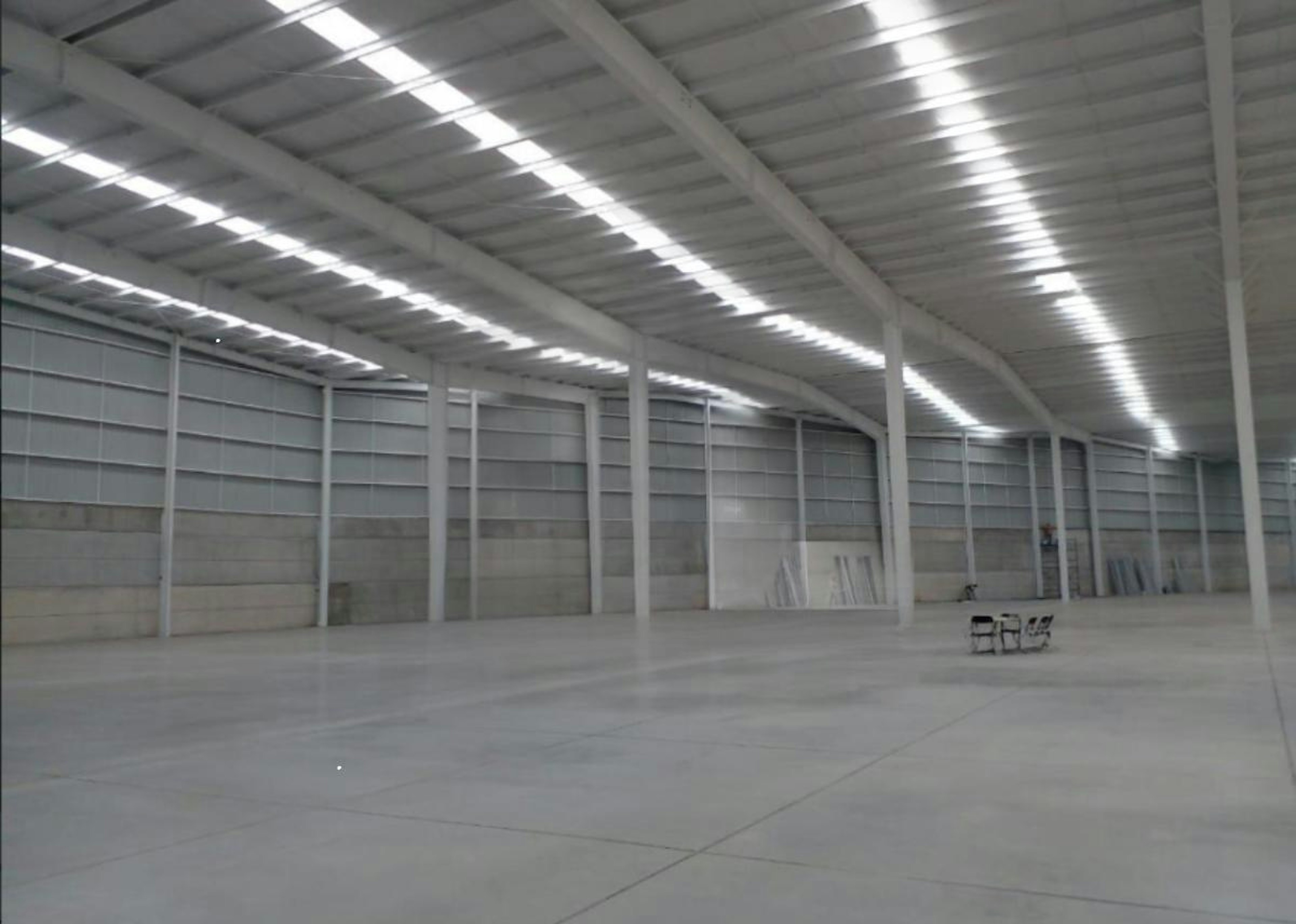 La imagen muestra un amplio espacio industrial de almacenamiento o logística. Es una nave industrial con techo alto y luminoso, paredes blancas y suelo de hormigón. El espacio está vacío excepto por algunas sillas y mesas plegables en un rincón. Se ve el logo de SIL Industry - Strategic Industry Logistics superpuesto en la imagen, sugiriendo que es una instalación de logística estratégica industrial.