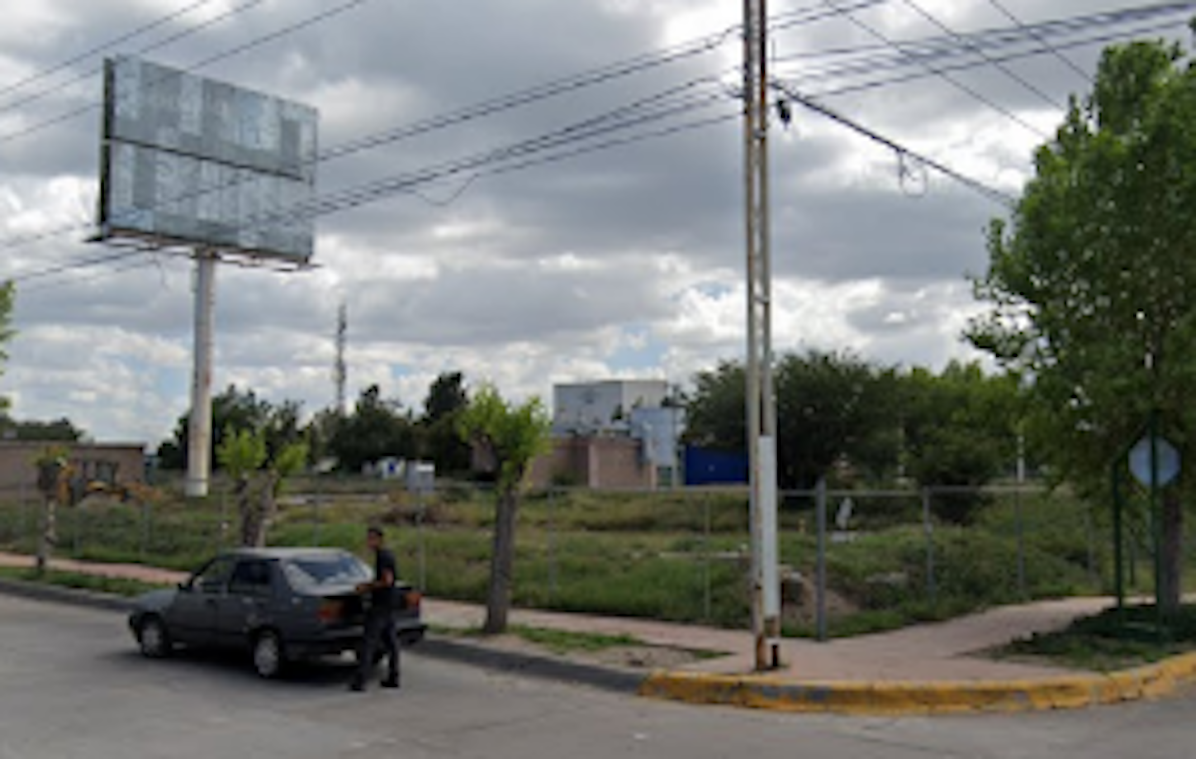 La imagen muestra un terreno baldío en una zona urbana, con un gran cartel publicitario vacío. Se observa vegetación dispersa, postes de electricidad y un vehículo estacionado. El área parece estar en desarrollo o transición, con potencial para futuros proyectos inmobiliarios. El cielo nublado y la infraestructura básica sugieren una zona en crecimiento.