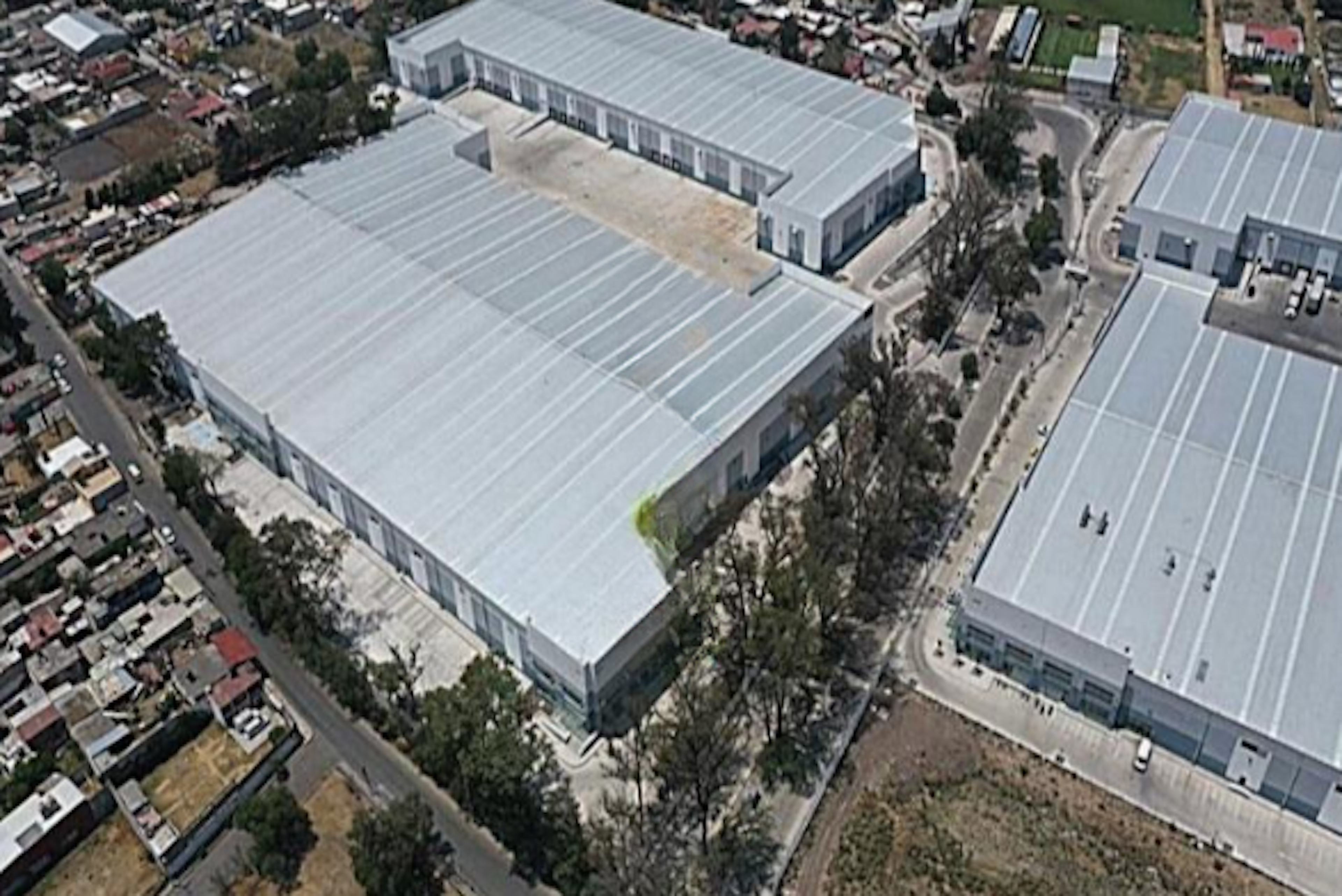 La imagen muestra una vista aérea de un complejo industrial o de almacenamiento. Se observan grandes edificios con techos blancos y rectangulares, probablemente naves industriales o bodegas. El complejo está rodeado por áreas residenciales y algunas zonas verdes. La ubicación parece ser una zona mixta industrial y residencial, con amplias instalaciones que sugieren actividades de almacenamiento, manufactura o distribución a gran escala.