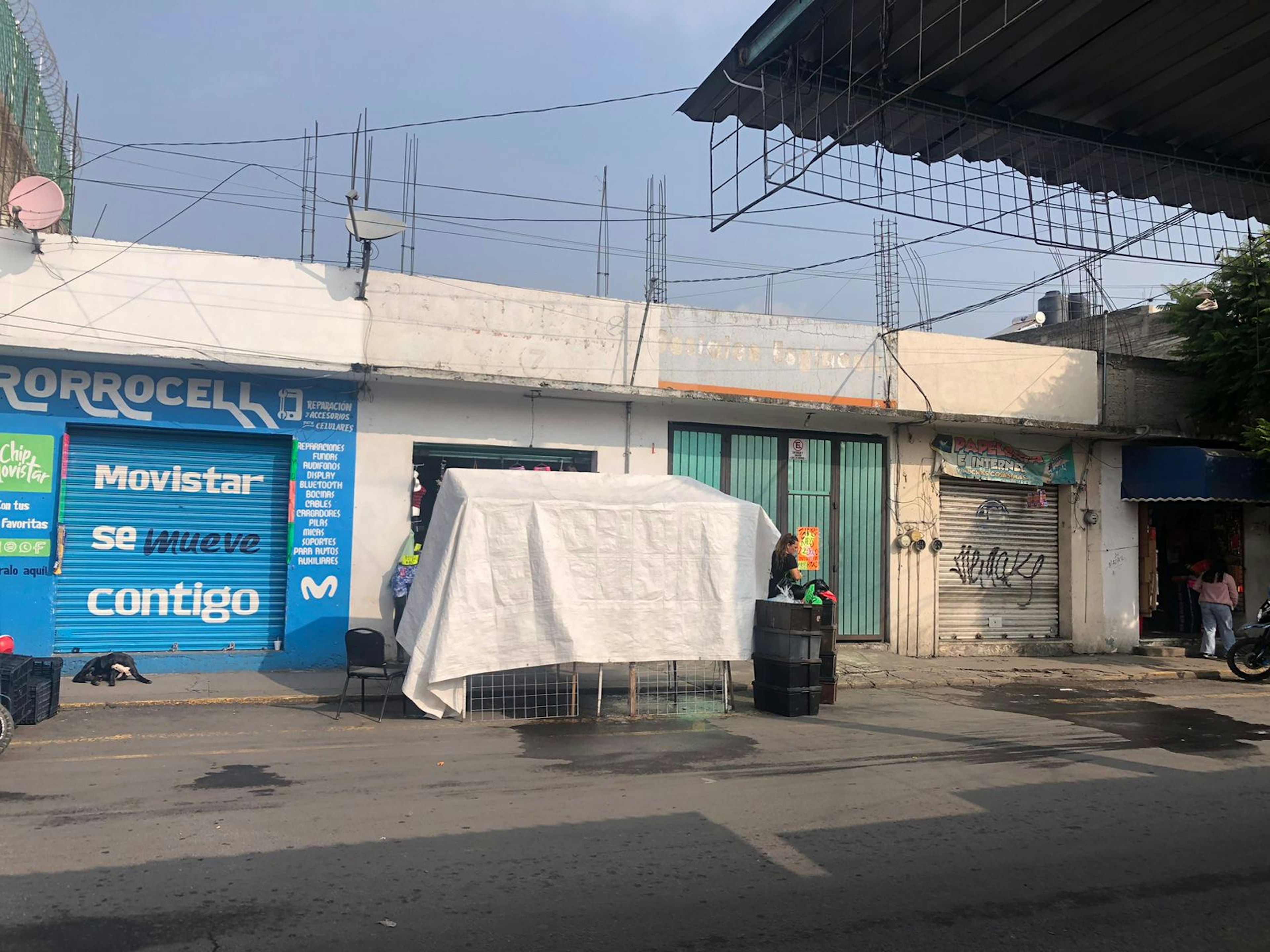 La imagen muestra una calle comercial con varias tiendas cerradas y una abierta, con un toldo blanco en la acera. La infraestructura parece antigua y necesita mantenimiento.