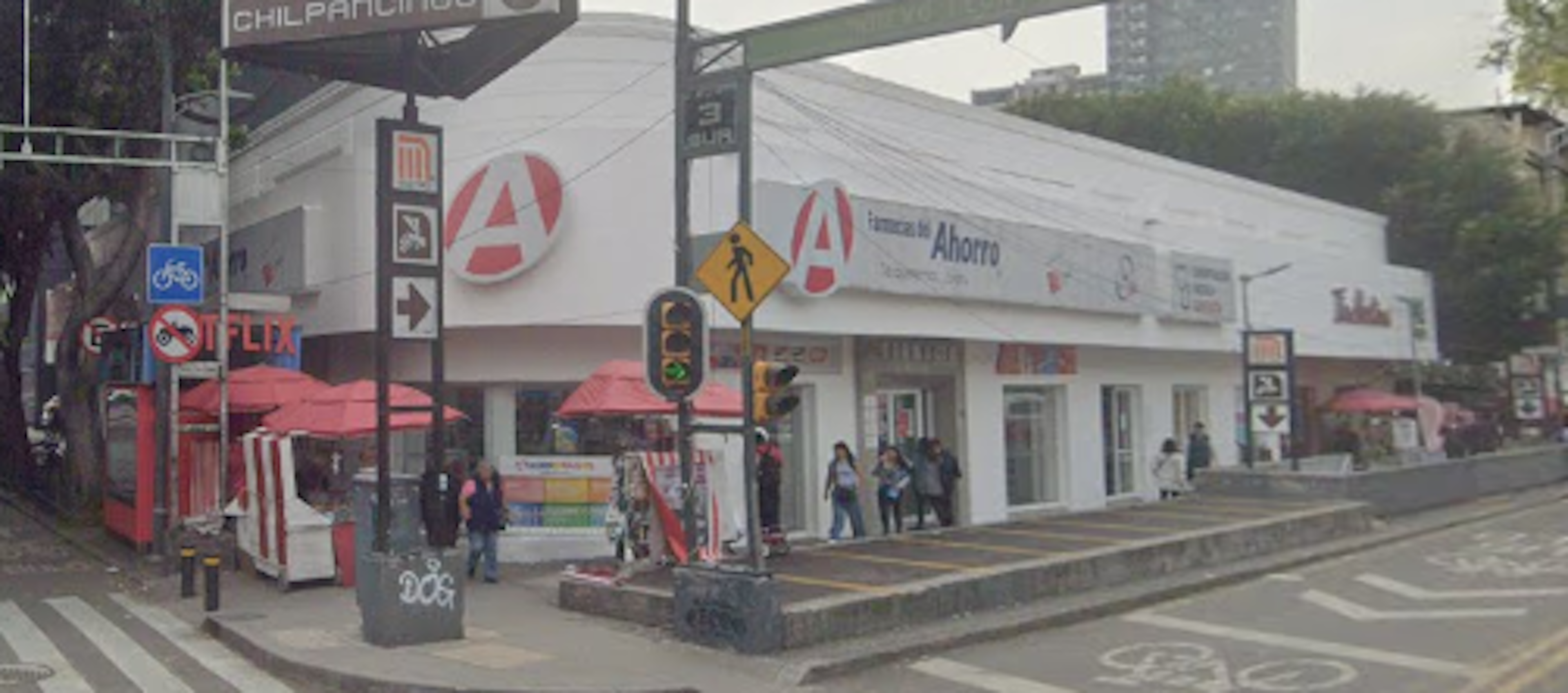 La imagen muestra una escena urbana con un edificio comercial blanco en la esquina, que alberga una tienda o banco llamado Ahorro. Hay señales de tráfico, semáforos y un cruce peatonal frente al edificio. Se observan algunos puestos ambulantes con sombrillas rojas en la acera. La zona parece ser un área comercial activa con peatones y actividad en la calle. El entorno sugiere una ubicación céntrica con potencial para negocios o inversiones inmobiliarias.