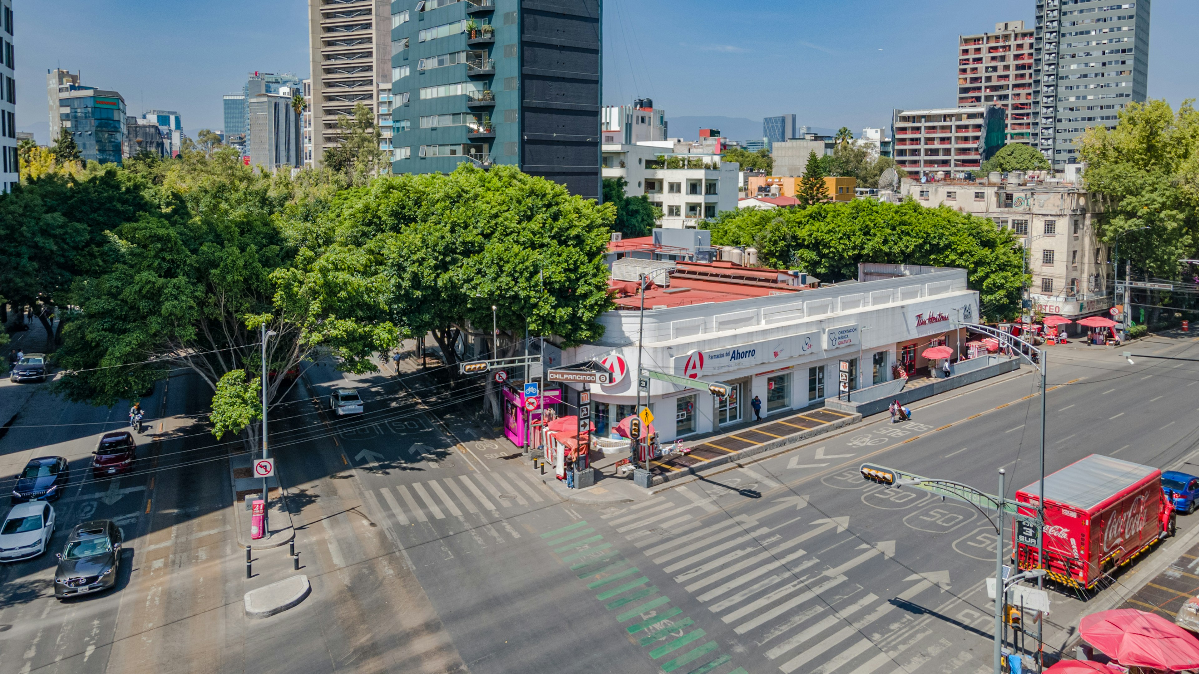 La imagen muestra una vista aérea de una zona urbana moderna con altos edificios de oficinas y apartamentos. En primer plano se aprecia una intersección con un pequeño centro comercial, rodeado de árboles y calles transitadas. La escena combina desarrollo urbano con áreas verdes, típico de un distrito comercial y residencial en una gran ciudad.