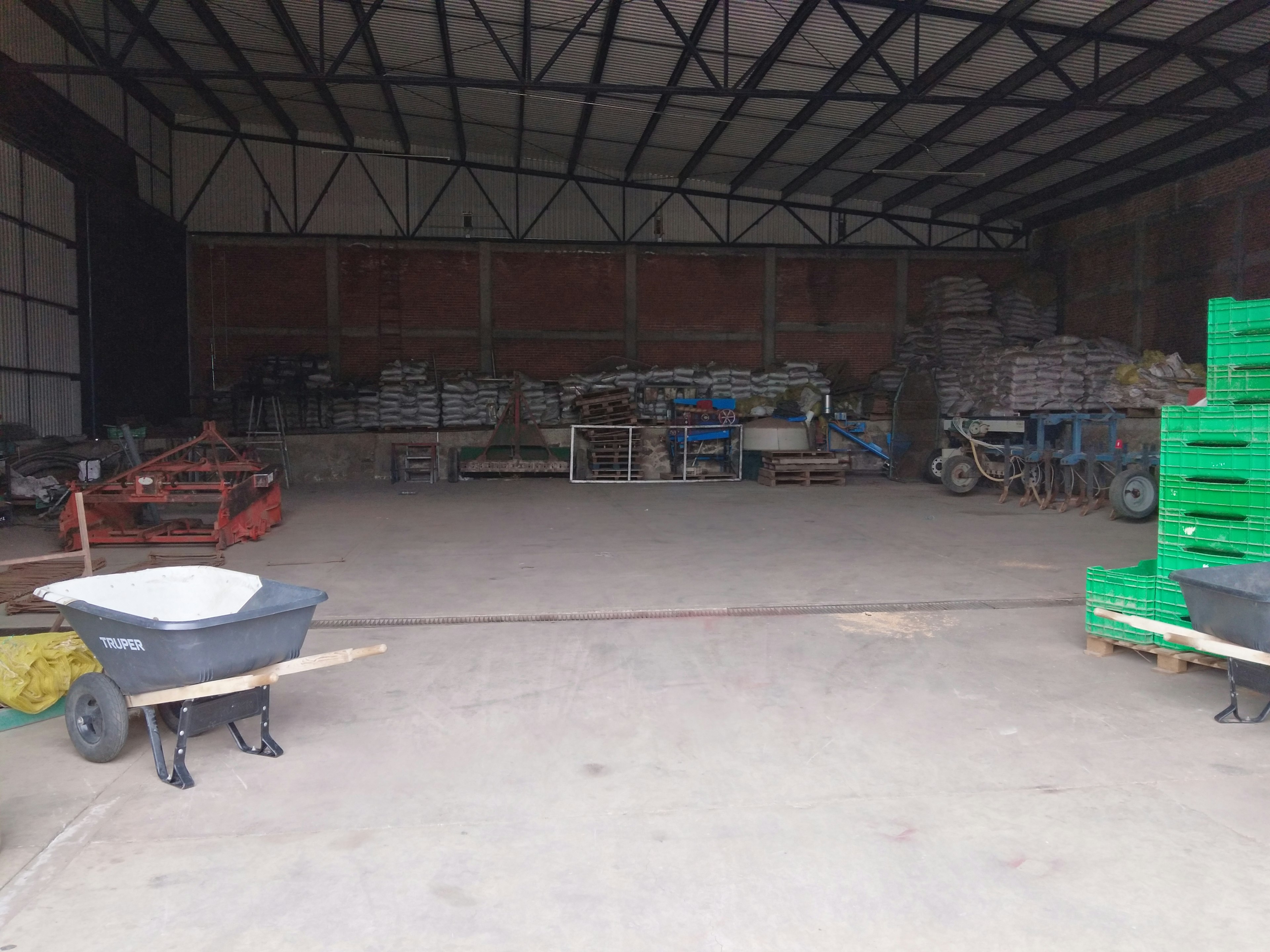 Esta imagen muestra un amplio almacén o bodega industrial con techo alto de metal. El espacio está lleno de equipos agrícolas, sacos apilados (posiblemente fertilizantes o semillas), cajas verdes de plástico y una carretilla en primer plano. El piso es de concreto y las paredes son de ladrillo rojo. El lugar parece ser utilizado para almacenamiento y operaciones agrícolas.