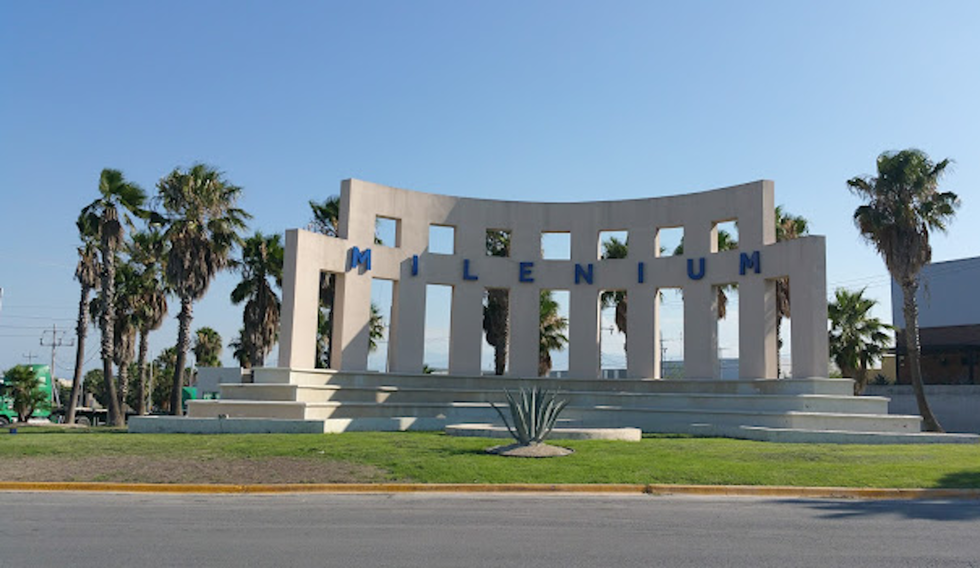 La imagen muestra un monumento arquitectónico moderno con el nombre MILENIUM en letras azules. Es una estructura curva de color beige con múltiples aberturas, rodeada de palmeras y césped. El cielo es azul claro, sugiriendo un clima cálido. Este tipo de estructura podría ser un punto de referencia en una zona residencial o turística, potencialmente aumentando el atractivo y valor de las propiedades cercanas.