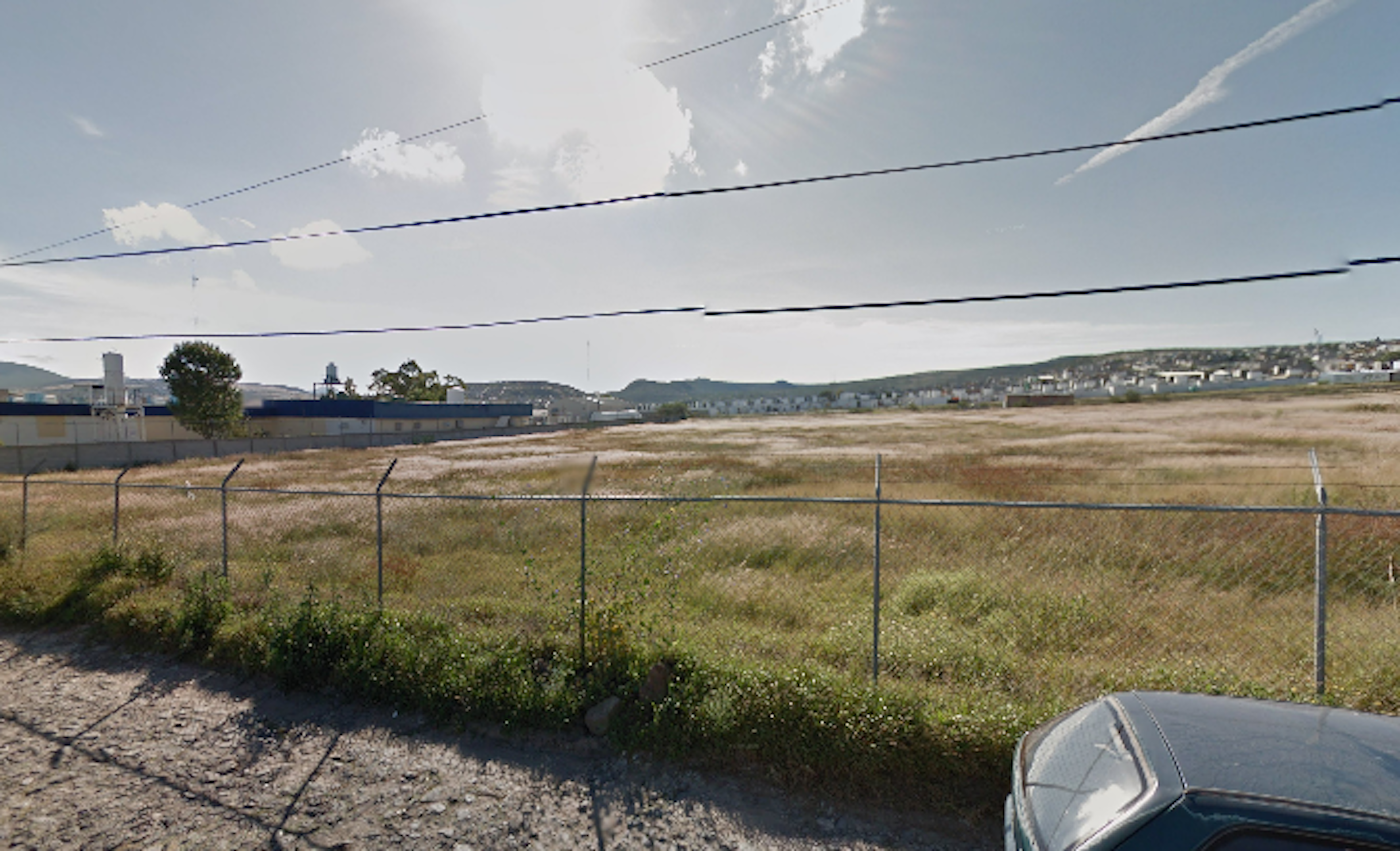 La imagen muestra un terreno baldío extenso y sin desarrollar, rodeado por una cerca de alambre. En el fondo se observan edificios industriales y una zona residencial, indicando que el área está en una ubicación semi-urbana. El terreno parece seco y cubierto de vegetación baja, sugiriendo potencial para desarrollo inmobiliario. Hay líneas eléctricas visibles y el cielo está parcialmente nublado, dando una sensación de amplitud al paisaje.