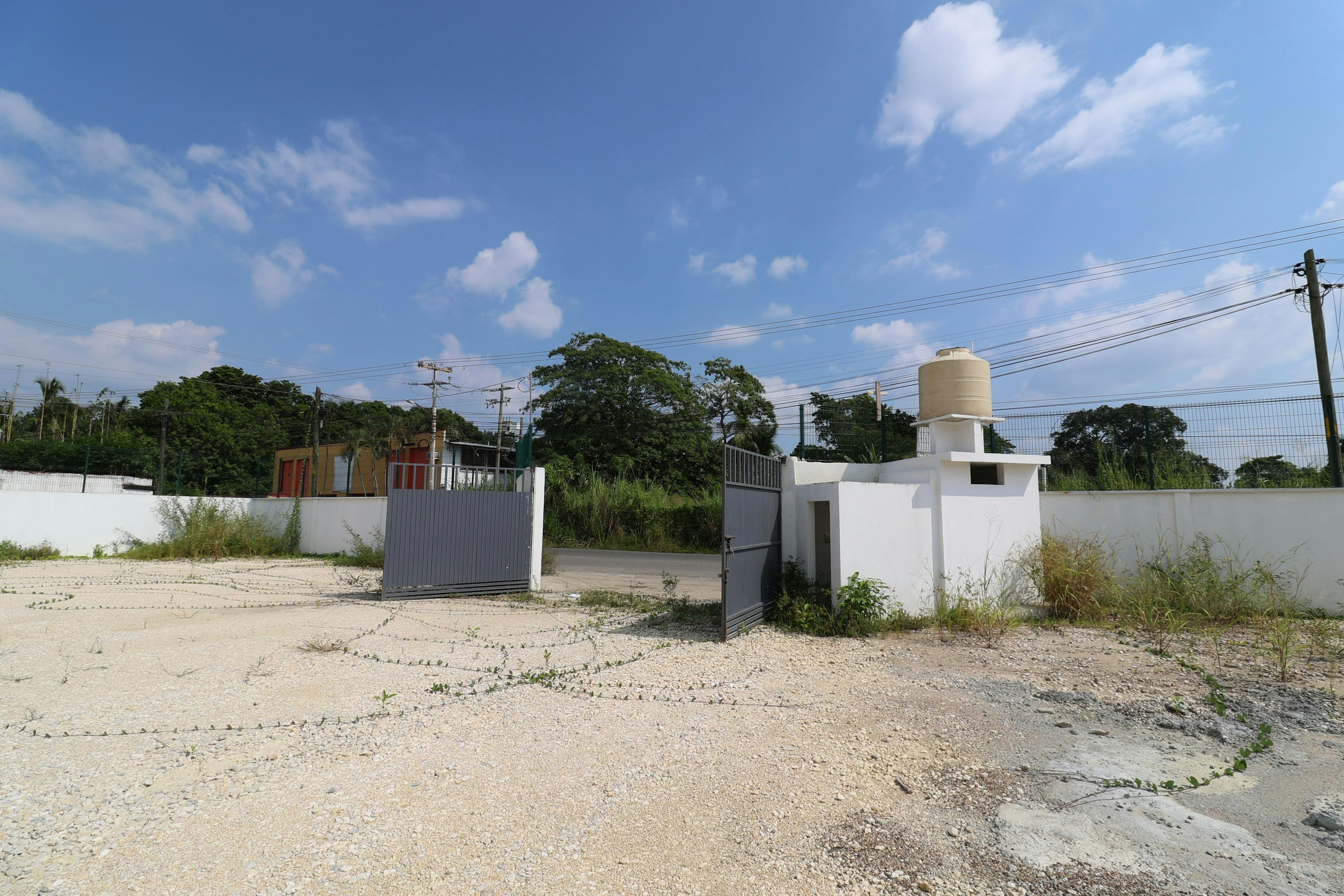 La imagen muestra un terreno baldío con una entrada cercada. Se observa una puerta metálica gris y una estructura blanca con un tanque de agua en la parte superior. El entorno sugiere una zona en desarrollo, con vegetación y postes eléctricos visibles. El cielo es azul con nubes dispersas, indicando un clima agradable. El terreno parece estar en una zona suburbana o rural en proceso de urbanización.