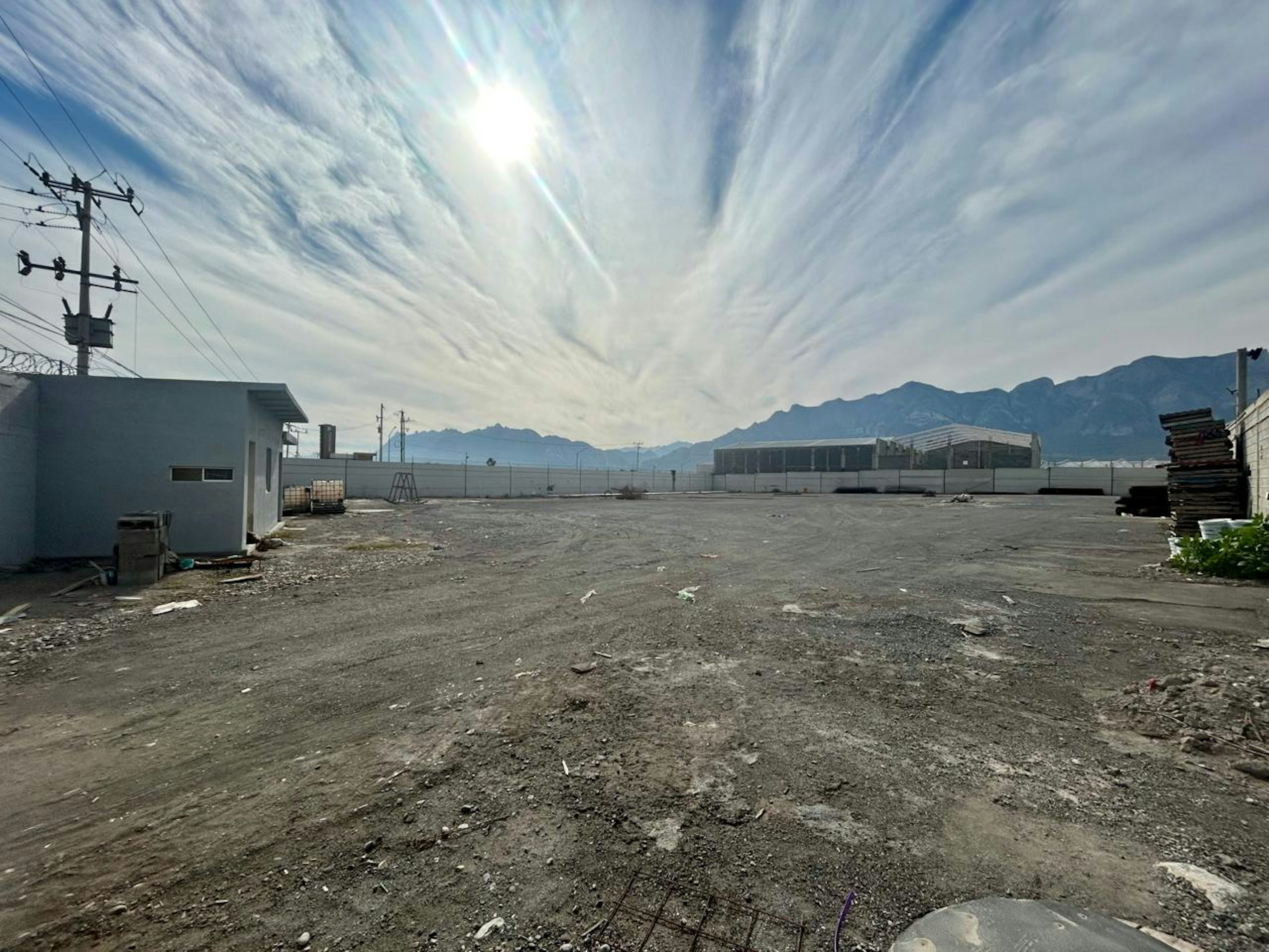 La imagen muestra un terreno baldío en una zona industrial o suburbana. Se ve un amplio espacio abierto y sin desarrollar, con suelo de tierra y algunos escombros. Al fondo se aprecian montañas y un cielo dramático con nubes. Hay algunas estructuras simples, postes eléctricos y un edificio blanco pequeño. El área parece estar en una etapa temprana de desarrollo o abandono, con potencial para construcción futura.