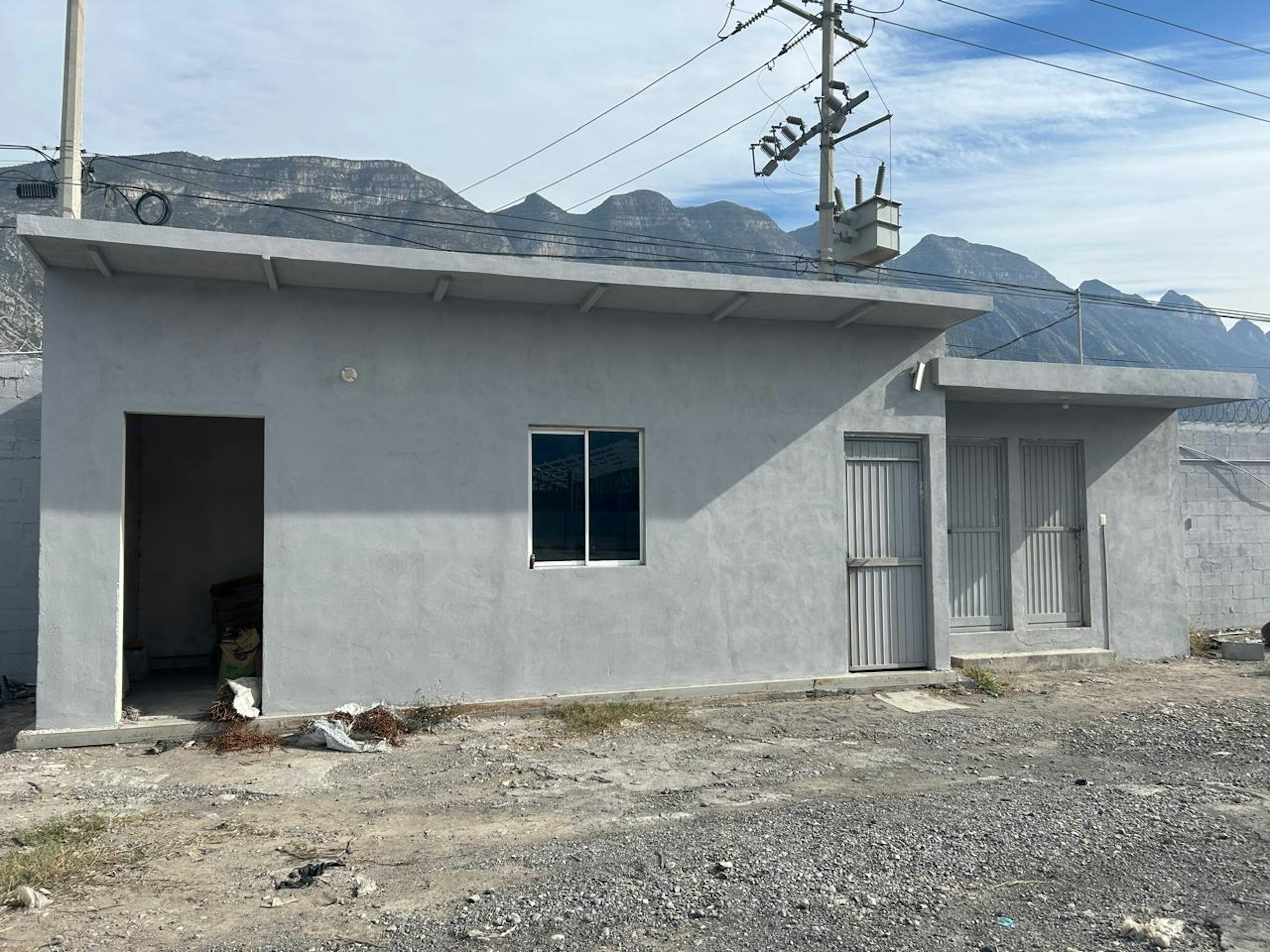 La imagen muestra una casa sencilla de una planta con paredes de concreto gris. Tiene una puerta metálica, una ventana y un techo plano. El entorno es rural, con montañas visibles al fondo. La propiedad parece estar en construcción o renovación, con terreno sin desarrollar alrededor. Se observan cables eléctricos y postes cerca de la casa, indicando acceso a servicios básicos.