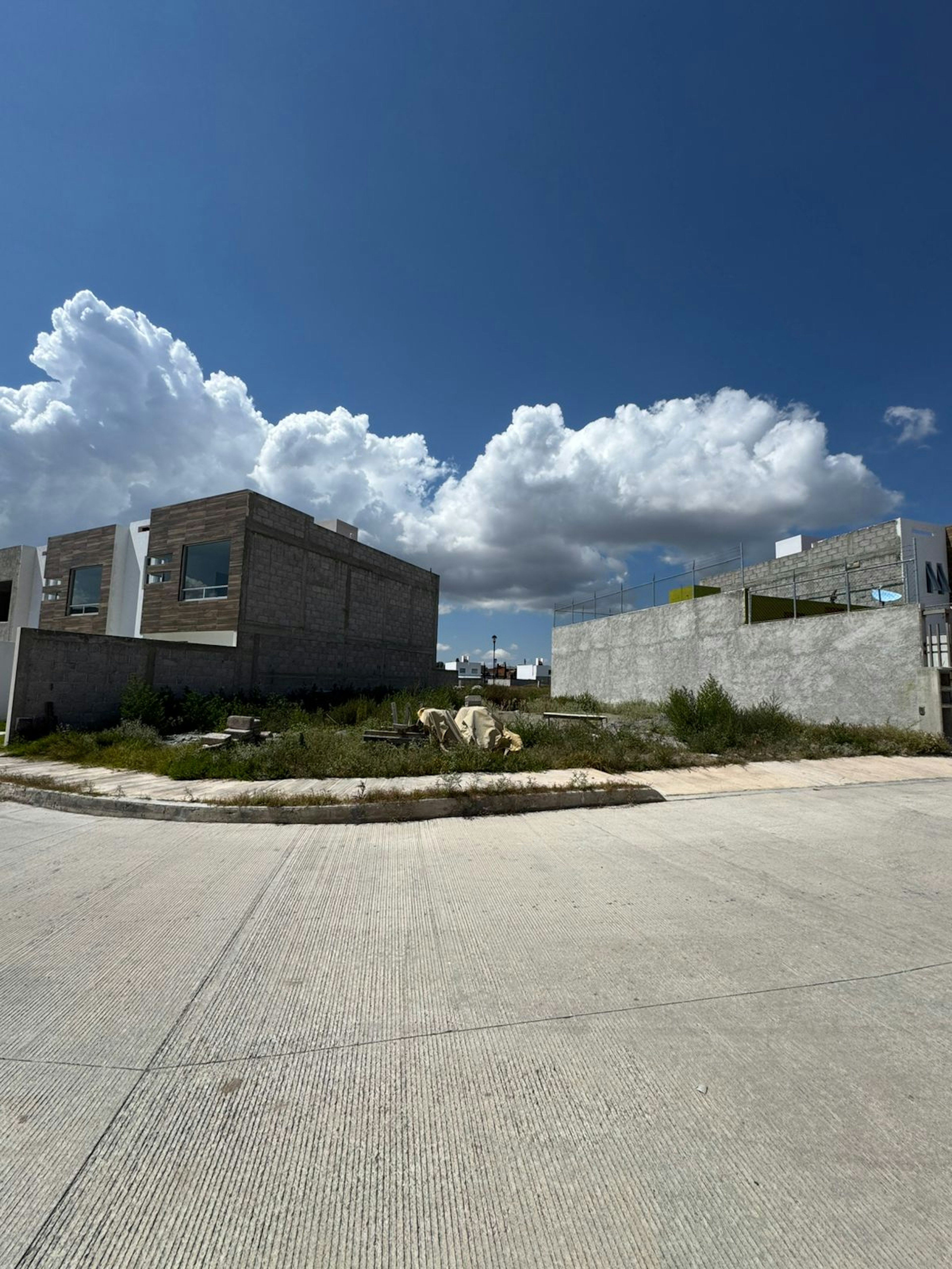 La imagen muestra un área urbana en desarrollo con edificios de construcción reciente y terrenos baldíos. Se observan estructuras de bloques de concreto, algunas parcialmente terminadas. Un cielo azul con nubes blancas domina la escena. En primer plano hay una calle pavimentada y un lote vacío con vegetación silvestre. El ambiente sugiere una zona residencial o comercial en proceso de urbanización.