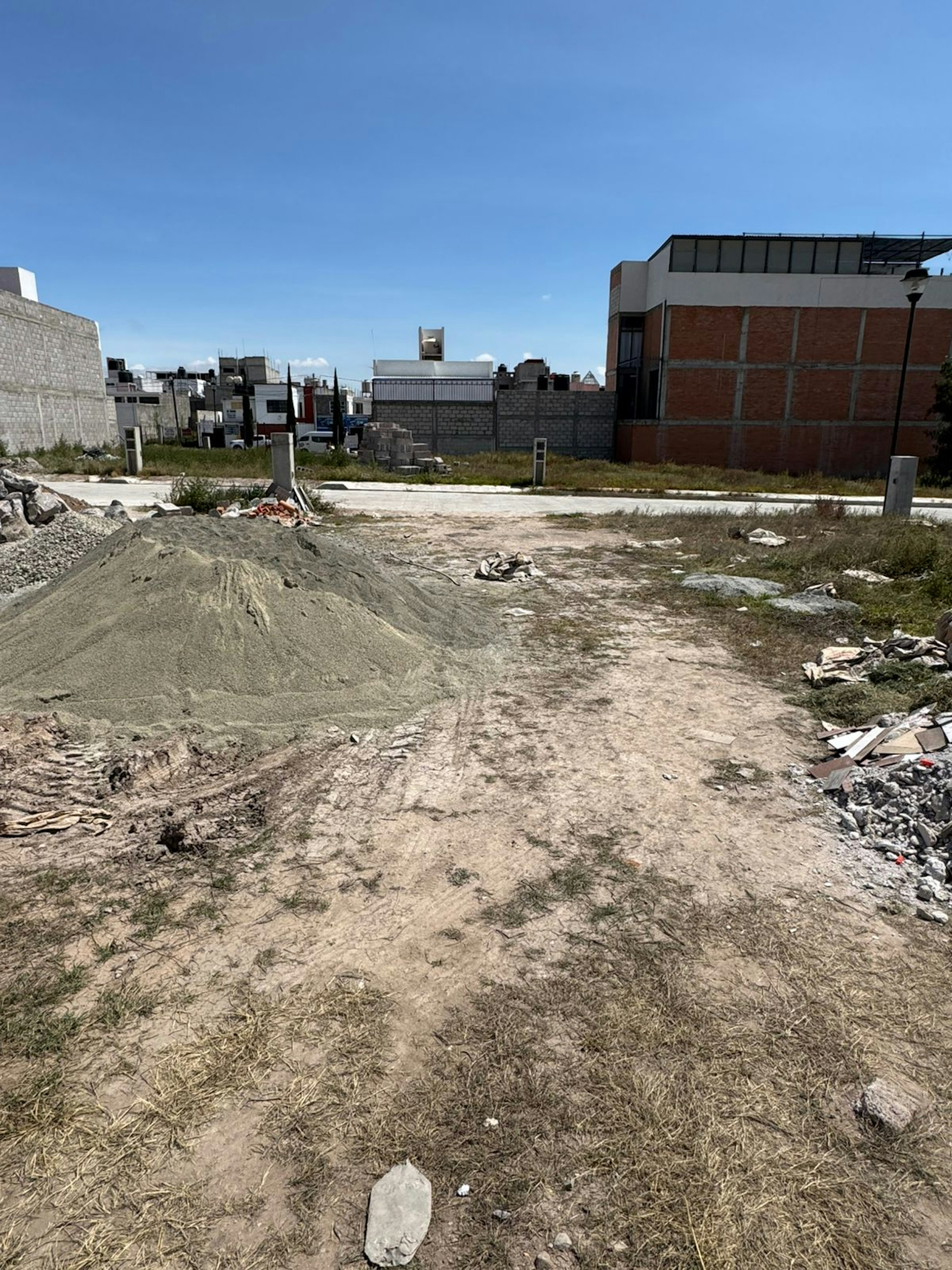 La imagen muestra un terreno baldío en una zona urbana en desarrollo. Se observa un área de construcción con montículos de tierra, escombros y materiales dispersos. Al fondo se ven edificios de ladrillo y concreto en diferentes etapas de construcción. El terreno parece estar en preparación para futuras edificaciones, indicando una posible expansión inmobiliaria en el área.