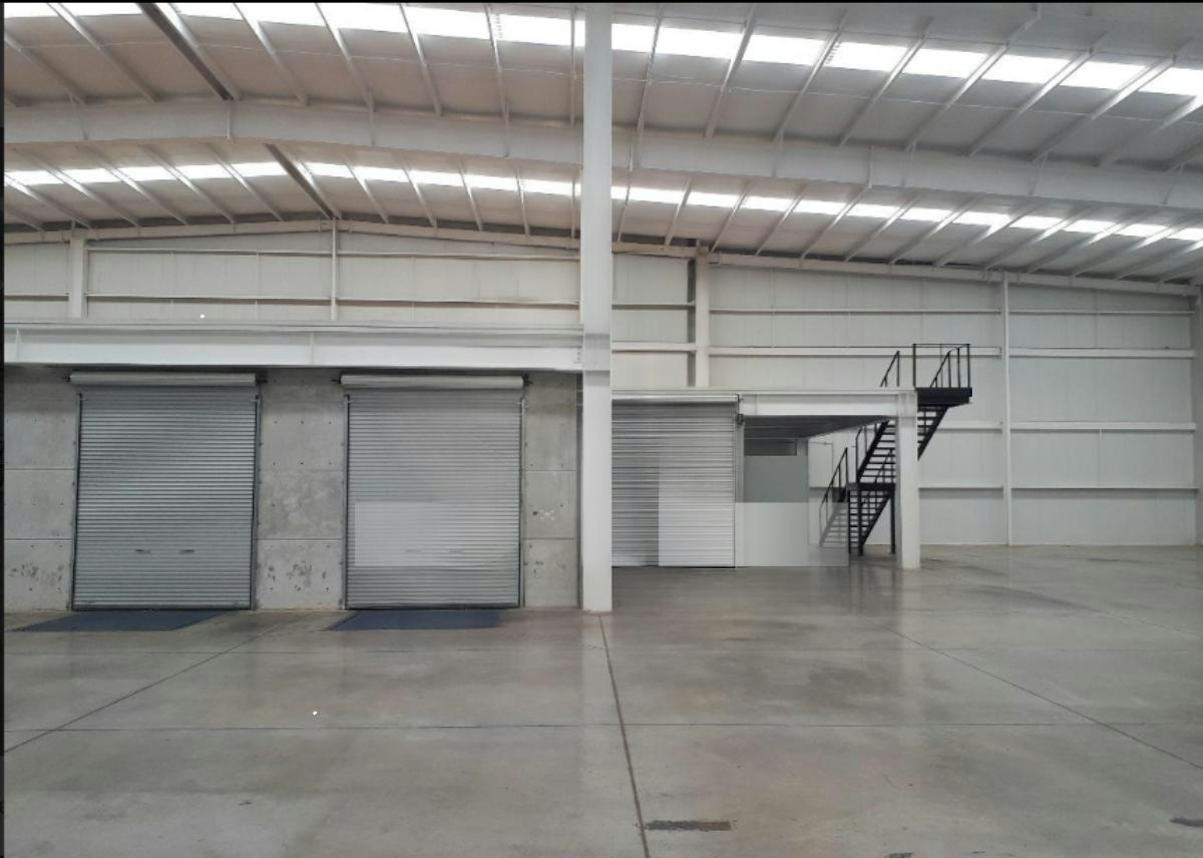 La imagen muestra el interior de una nave industrial o almacén logístico. Se observa un espacio amplio con techo alto y estructura metálica. Hay puertas enrollables cerradas y una escalera metálica que conduce a un nivel superior. El piso es de concreto y las paredes son blancas. En el suelo se aprecia el logotipo y texto de Strategic Industry Logistics Industry, sugiriendo que es un espacio para operaciones logísticas industriales.