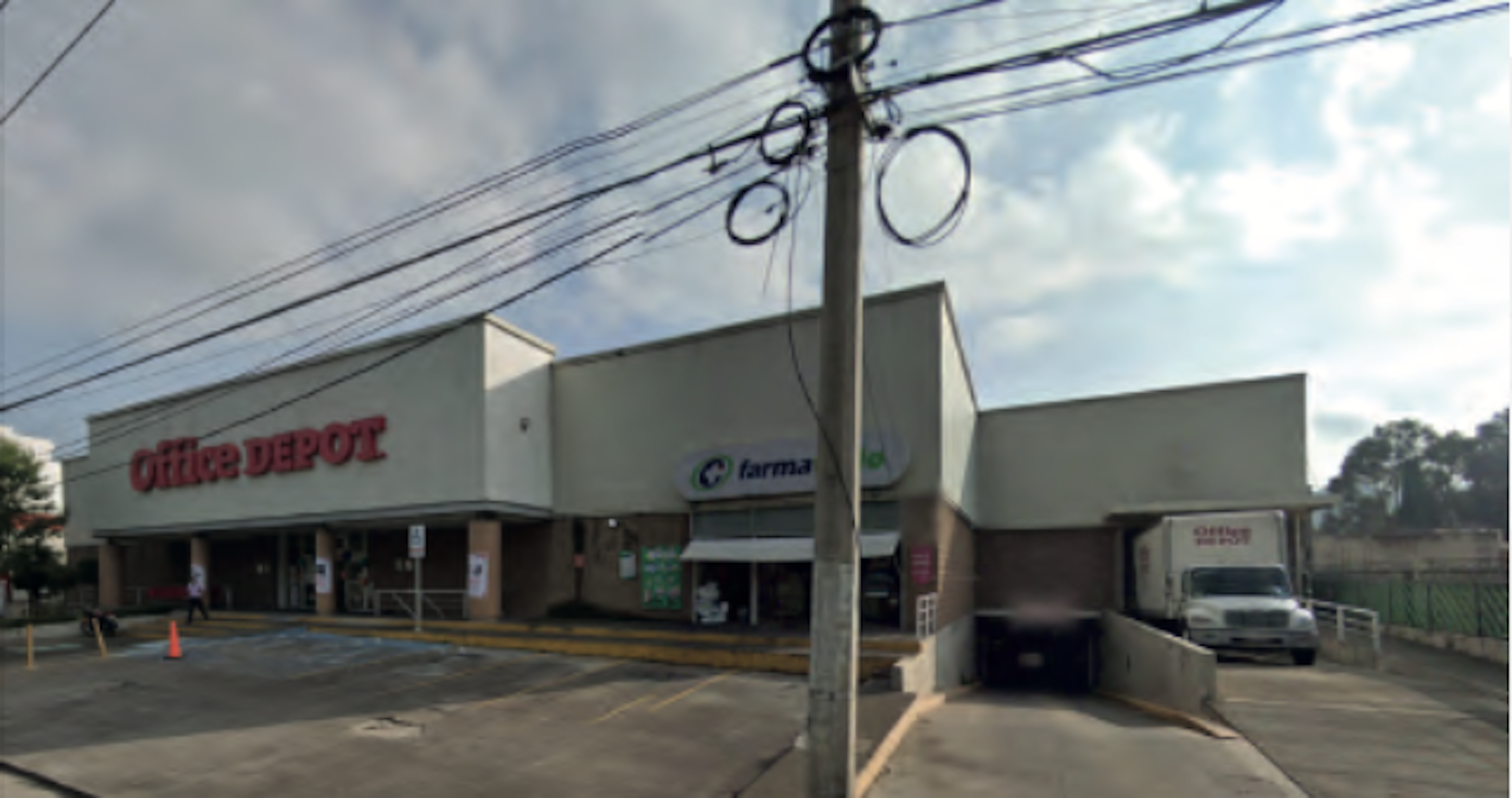 La imagen muestra un edificio comercial que alberga varias tiendas, incluyendo Office Depot y una farmacia. La estructura es de un solo piso con fachada blanca y base de ladrillo. Se observa un estacionamiento en frente y un camión de reparto en un área de carga. El edificio parece estar ubicado en una zona suburbana, con cables eléctricos visibles y un cielo nublado de fondo.