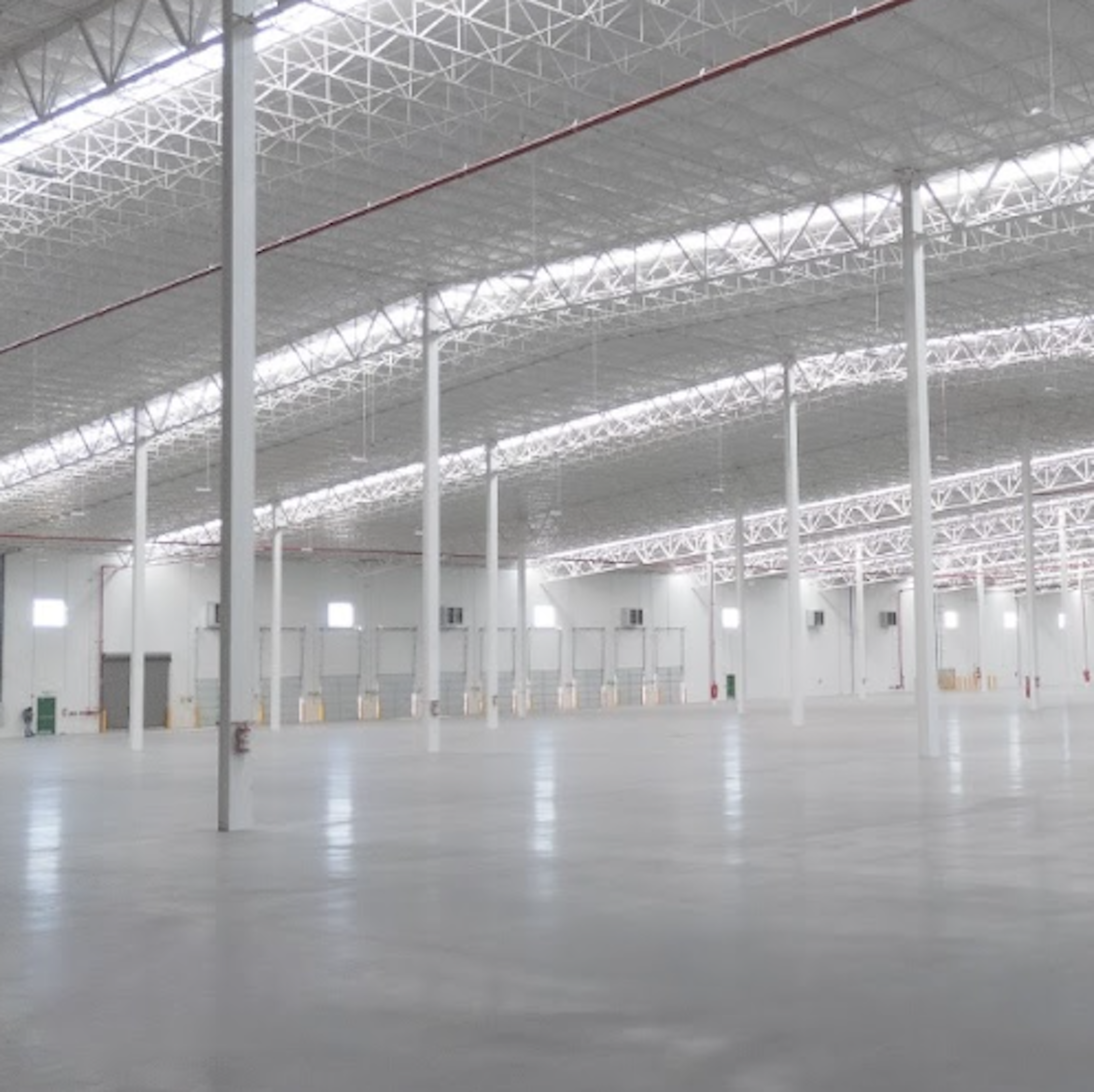 La imagen muestra un amplio espacio industrial o de almacenamiento con techo alto y estructura metálica. El interior es luminoso y espacioso, con columnas blancas que sostienen el techo. El suelo es de concreto pulido y hay varias puertas o entradas visibles a lo largo de las paredes. La nave está vacía, lo que sugiere que está lista para ser ocupada o utilizada para diversos fines comerciales o logísticos.