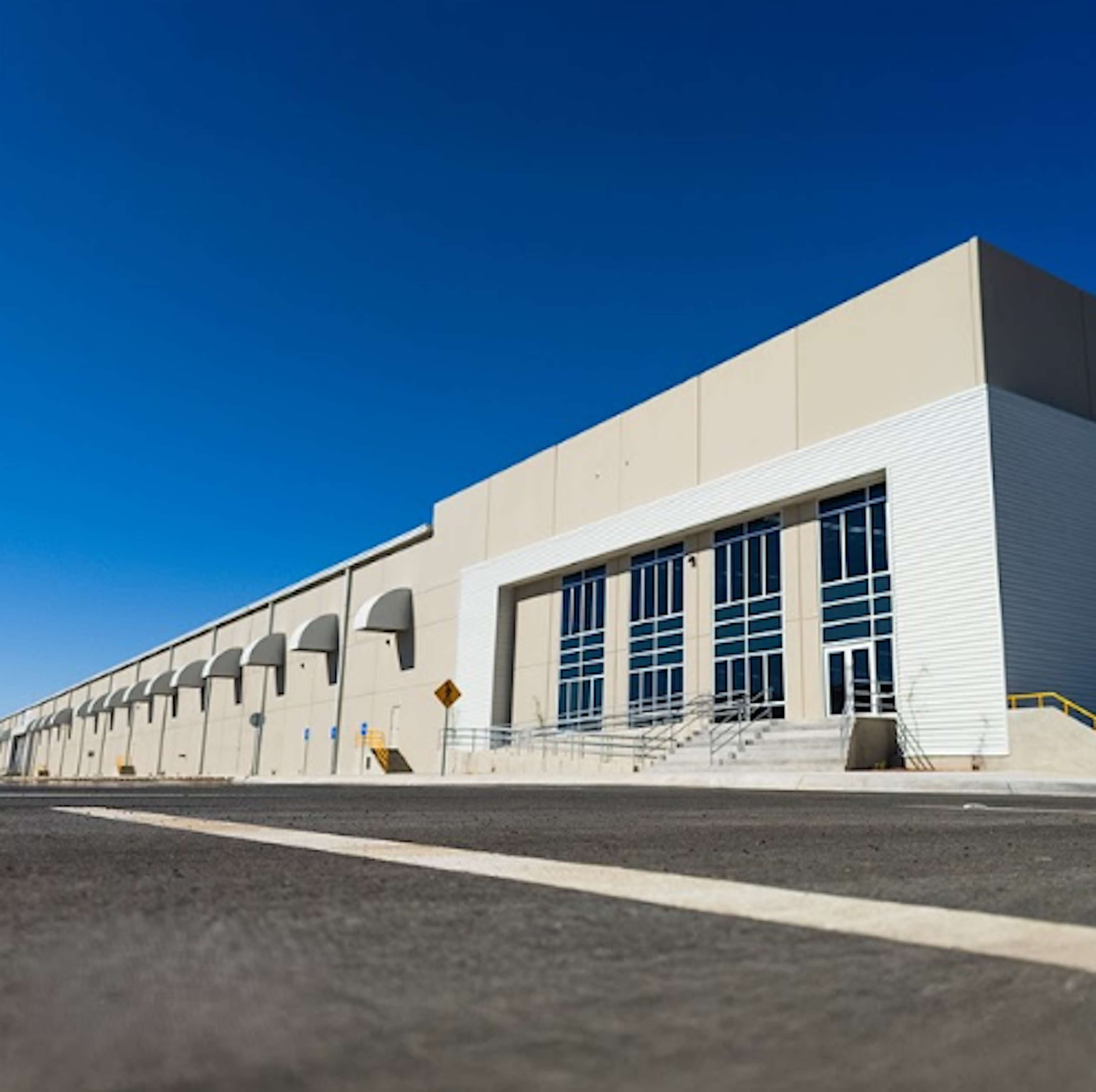 La imagen muestra un edificio comercial o industrial moderno con un diseño funcional. Tiene una fachada blanca y beige con grandes ventanales en la entrada principal. La estructura es extensa, con una serie de arcos decorativos a lo largo de un lado. El edificio parece ser un almacén o centro de distribución, con un amplio espacio de estacionamiento o área de carga visible. El cielo es de un azul intenso, lo que resalta la arquitectura limpia y moderna del inmueble.