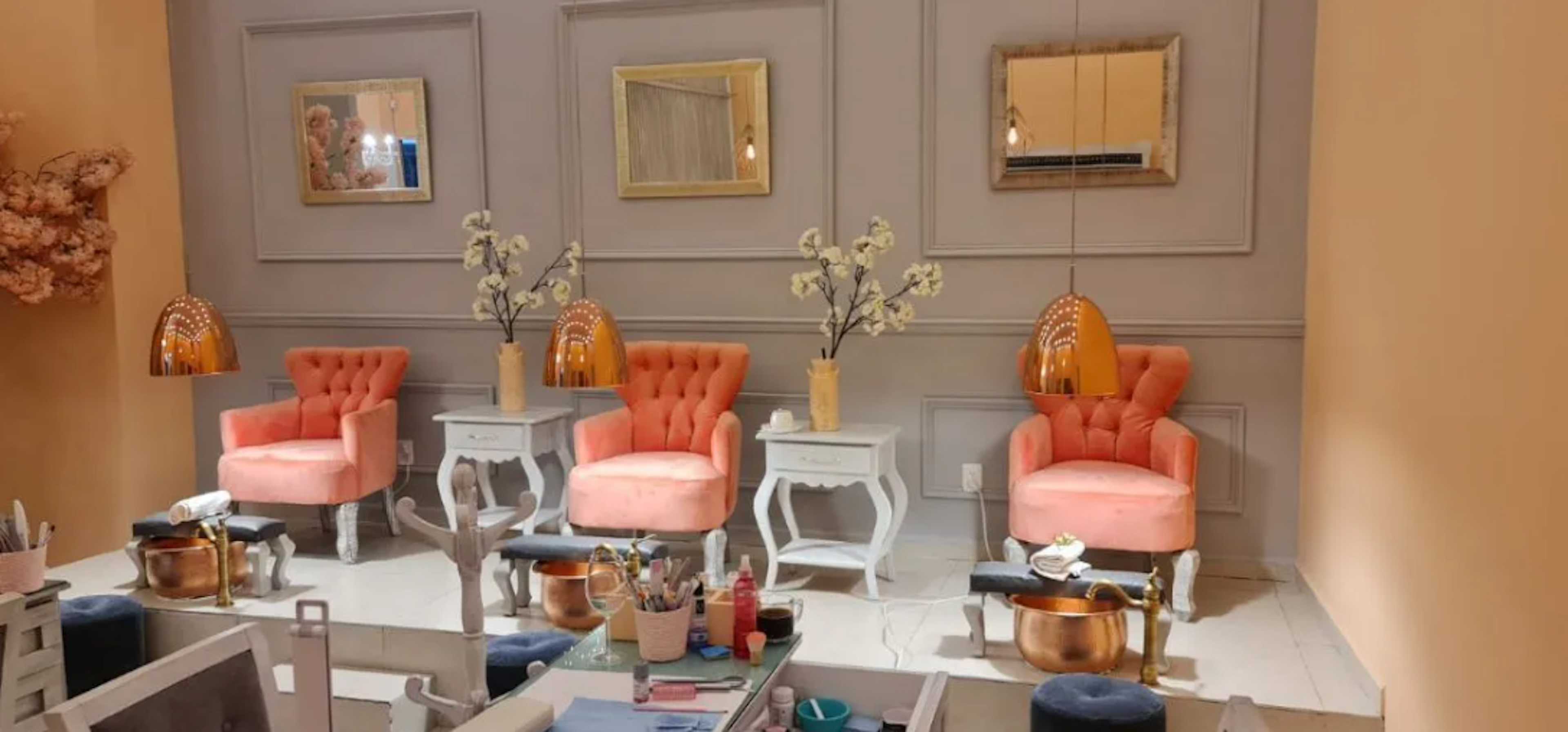 This image shows an elegantly designed beauty salon or spa interior. The space features three coral-colored tufted armchairs, each paired with a white ornate side table and a golden dome-shaped pendant light. The wall behind is decorated with panel moldings and framed mirrors. The foreground displays pedicure basins and beauty supplies, indicating a nail care service area. The color scheme is soft and feminine, with coral, gold, and white tones dominating the space.