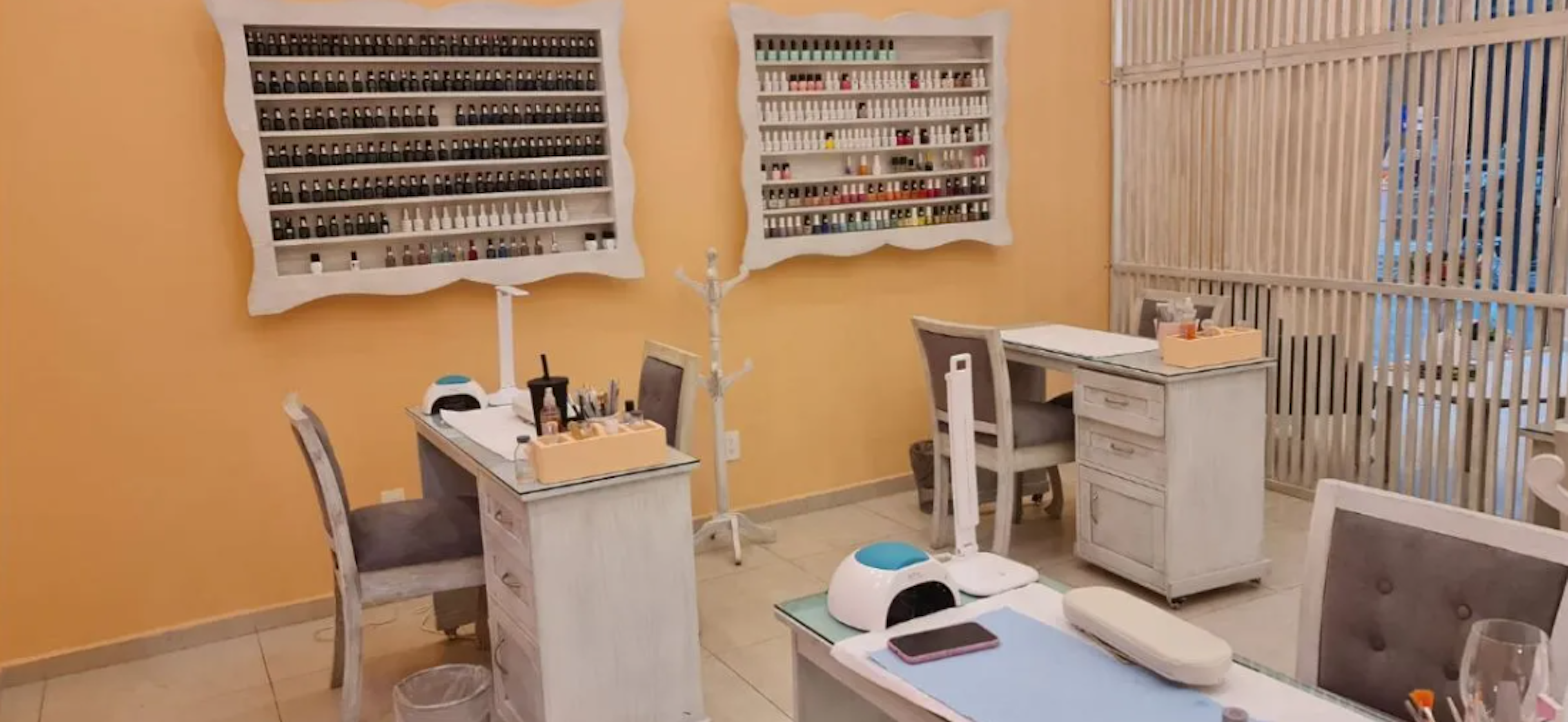 Esta imagen muestra un salón de manicura y pedicura bien equipado. Las paredes son de color amarillo cálido, con estantes blancos que exhiben una gran variedad de esmaltes de uñas. Hay varias estaciones de trabajo con mesas blancas y sillas grises. El espacio está organizado y limpio, con equipos modernos como lámparas UV visibles. La decoración es minimalista y funcional, creando un ambiente profesional y acogedor para los clientes.