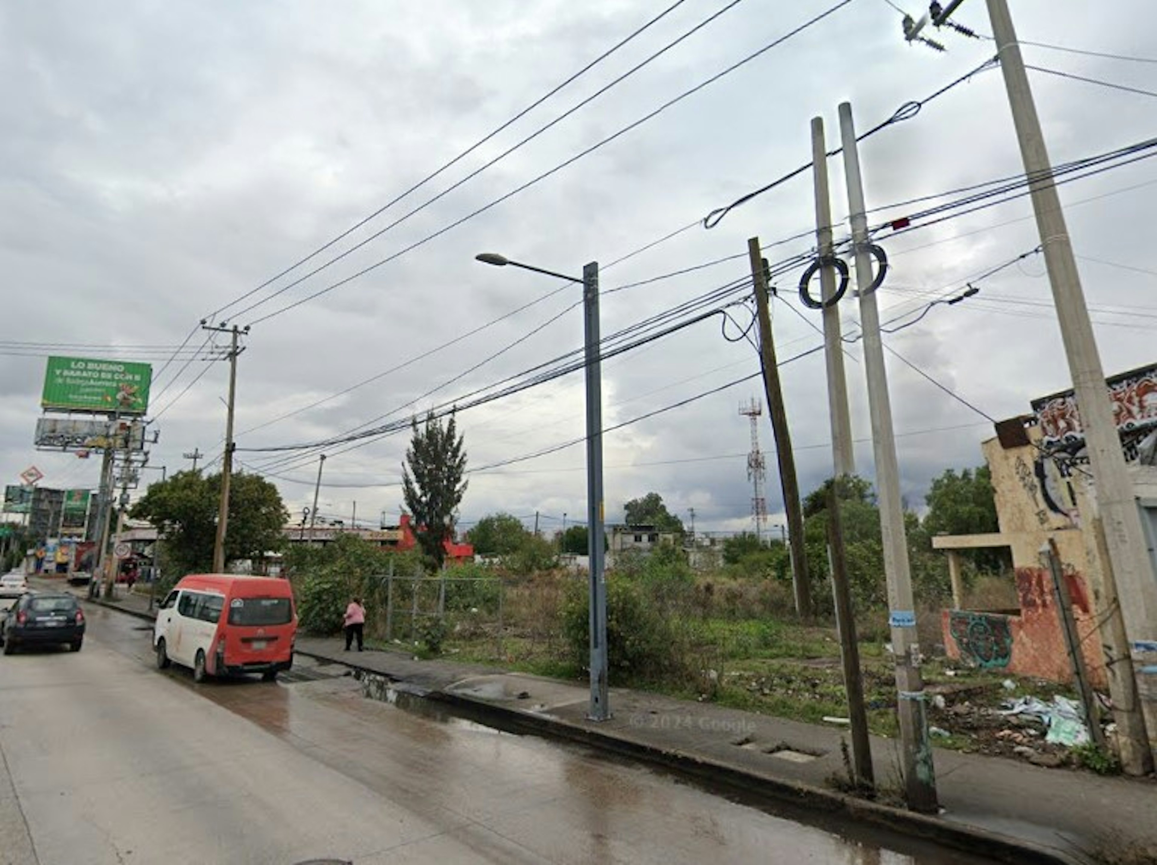 La imagen muestra una calle en una zona urbana con apariencia de suburbio o área en desarrollo. Se observan postes de electricidad, cables aéreos, un terreno baldío con vegetación, y algunas construcciones sencillas. La infraestructura parece básica, con una calle pavimentada pero sin aceras bien definidas. Hay vehículos estacionados, incluyendo una furgoneta roja. El cielo está nublado, sugiriendo un clima húmedo. El entorno general indica una zona de potencial desarrollo inmobiliario.