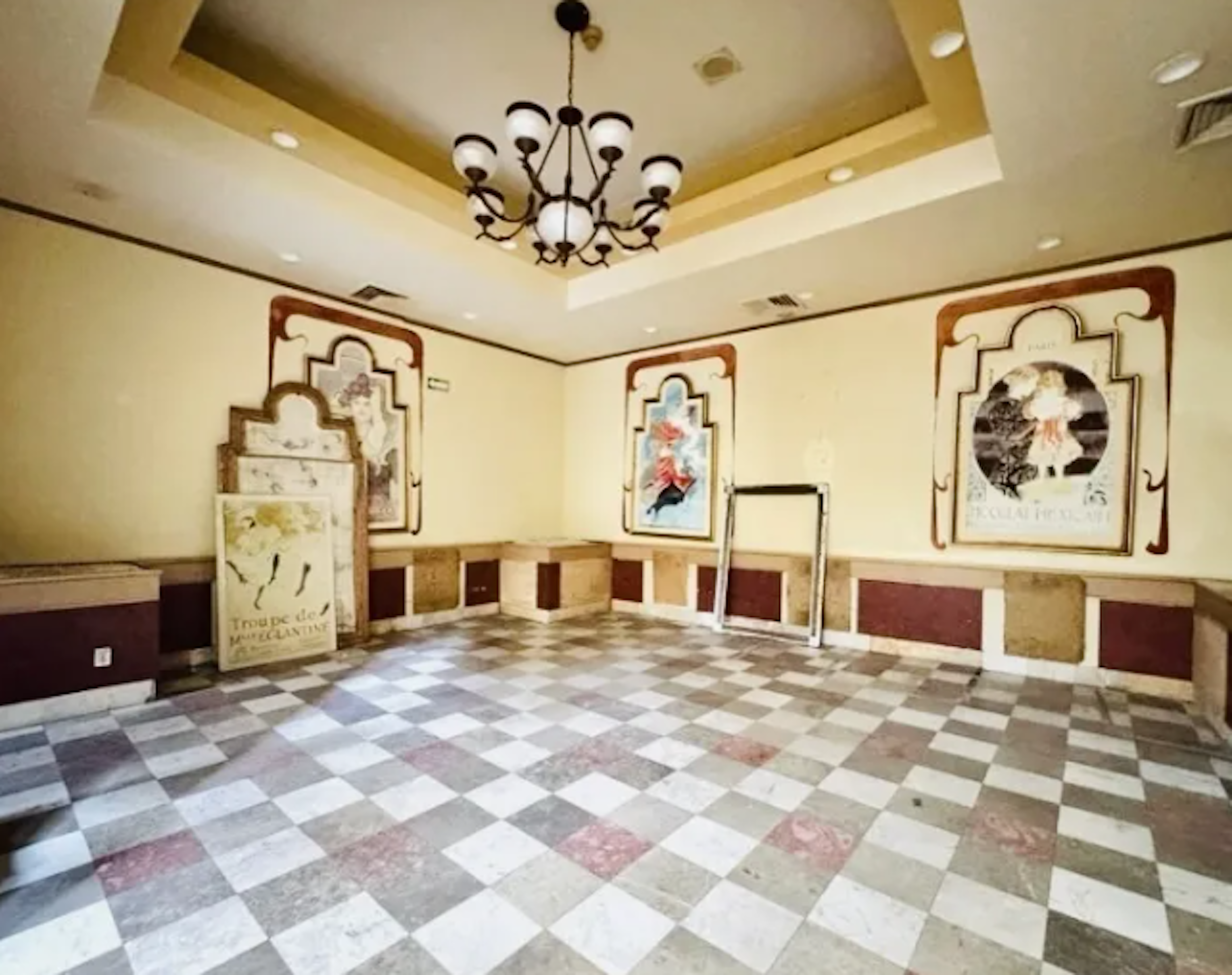 This image shows a spacious interior room with elegant decor. It features a checkered marble floor, ornate wall panels with artistic frames, and a grand chandelier. The room has a high ceiling with recessed lighting. The space appears to be part of a luxury property, possibly a hotel or upscale event venue. The decor combines classical and modern elements, creating a sophisticated atmosphere.