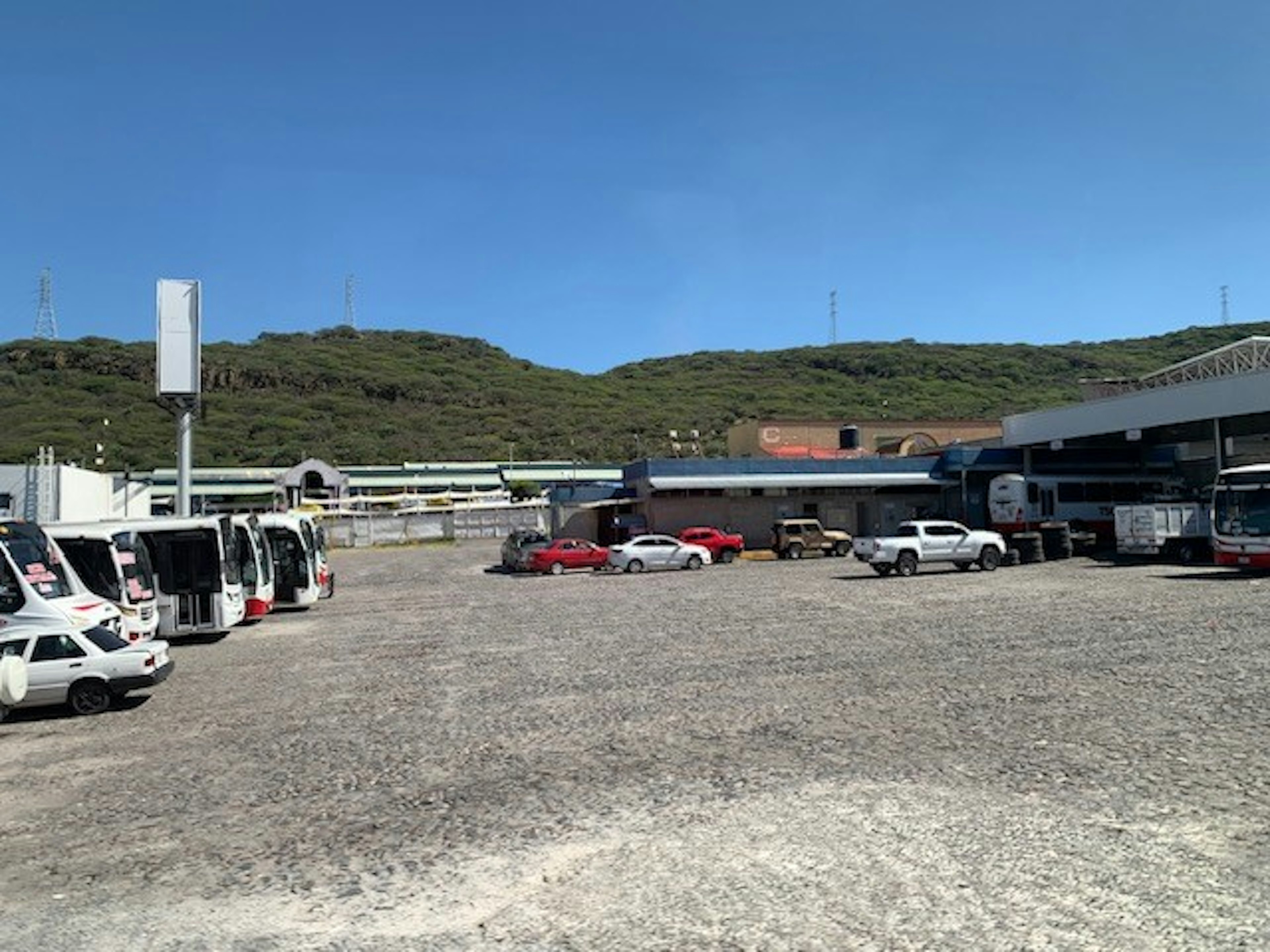 La imagen muestra un amplio estacionamiento o patio de maniobras en un área industrial o comercial. Se observan varios vehículos estacionados, incluyendo autobuses y automóviles. Al fondo se ven edificios y estructuras, probablemente almacenes o instalaciones comerciales. El terreno es de grava y está rodeado por colinas verdes. El cielo es azul y despejado, sugiriendo un clima favorable.