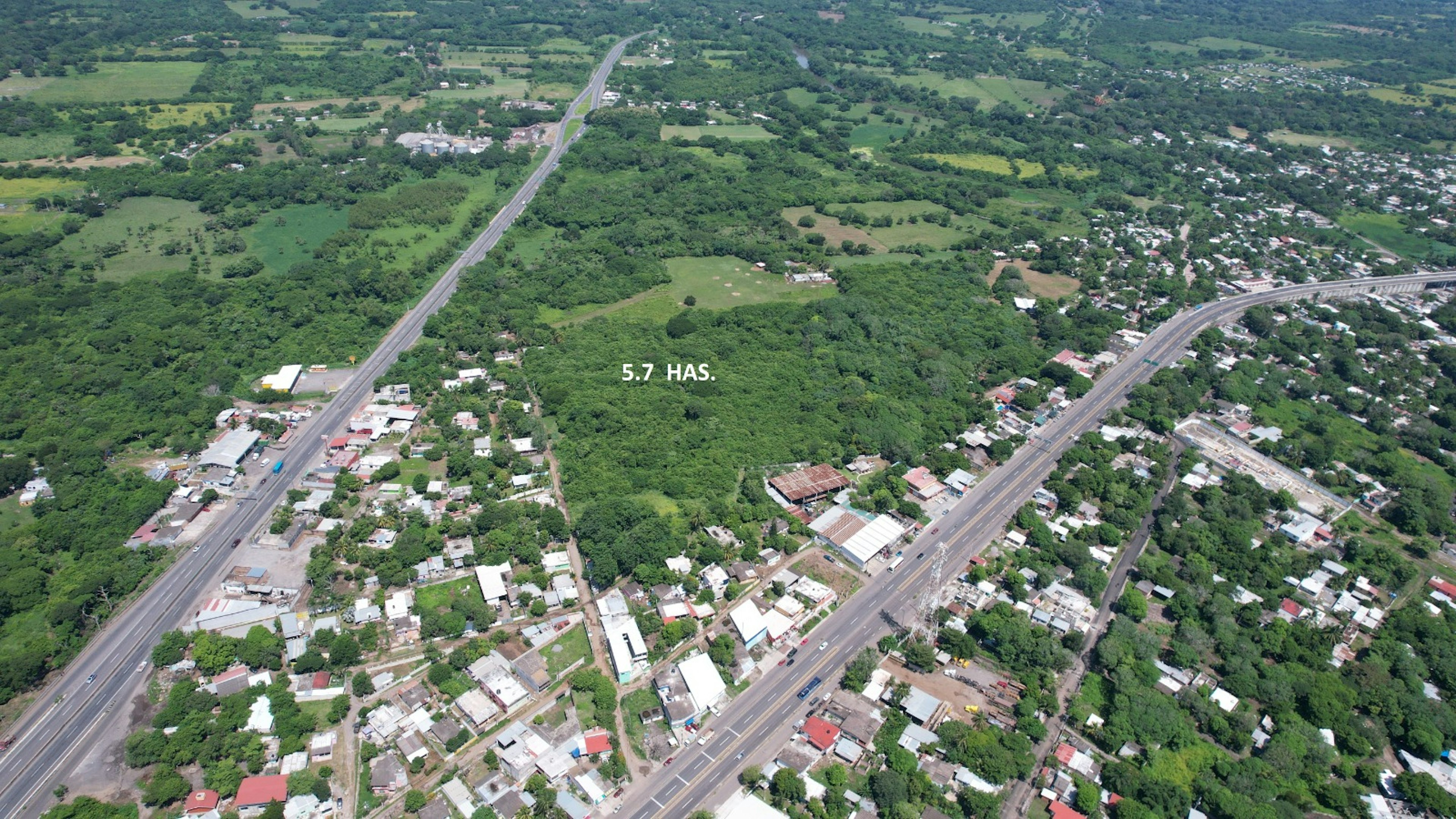 Esta imagen aérea muestra un área suburbana con una mezcla de zonas residenciales y espacios verdes. Se destacan dos carreteras principales que atraviesan la imagen. En el centro, se indica una parcela de 5.7 hectáreas, probablemente disponible para desarrollo inmobiliario. La zona parece tener potencial para expansión urbana, con una combinación de áreas construidas y terrenos sin desarrollar.