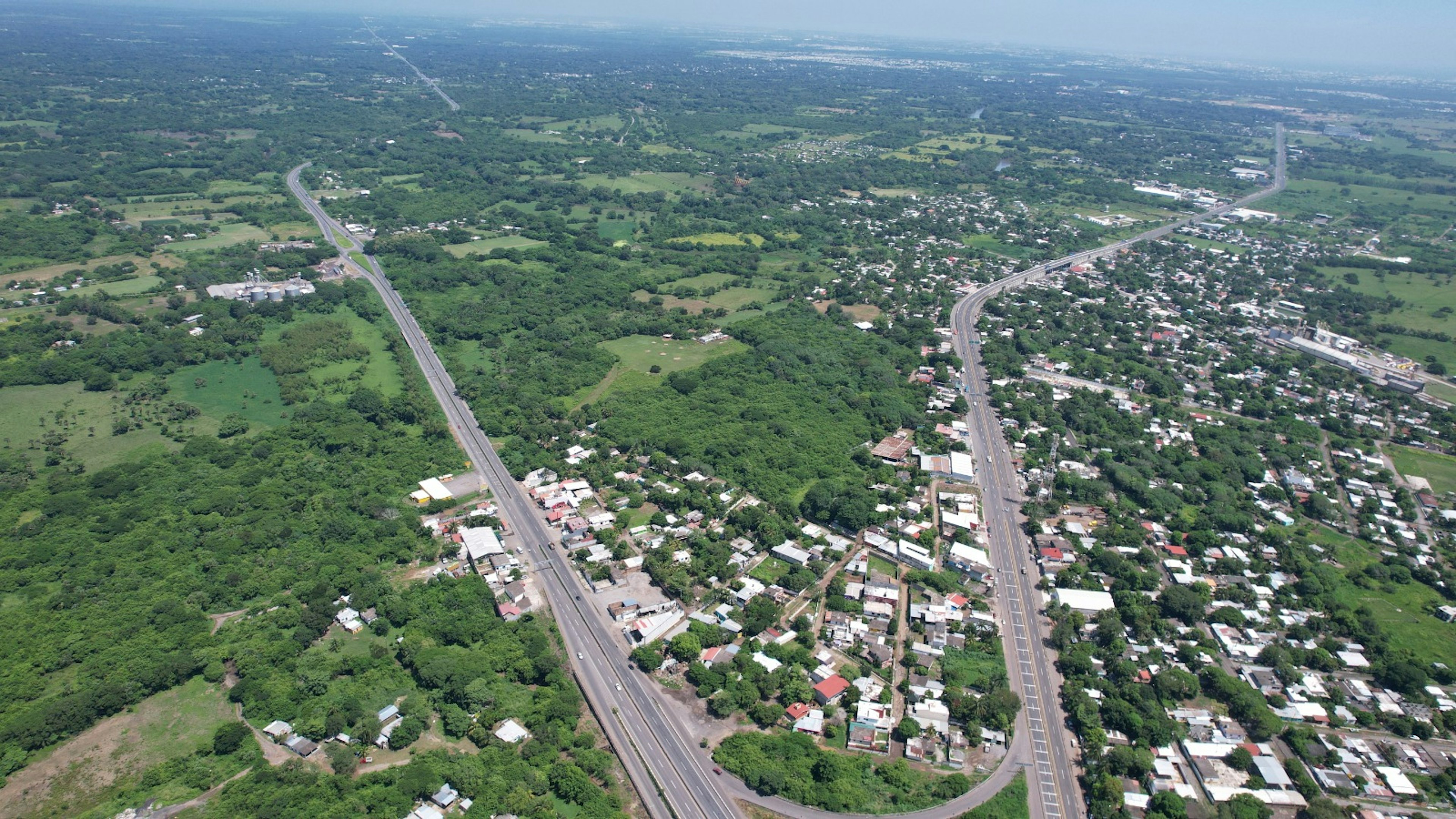 Esta imagen aérea muestra un área suburbana con una mezcla de zonas residenciales y espacios verdes. Se observan dos carreteras principales que atraviesan el paisaje, dividiendo el terreno en sectores. Las viviendas están distribuidas de manera dispersa entre la vegetación, lo que sugiere un desarrollo urbano de baja densidad. La presencia de amplias áreas verdes indica potencial para futuros desarrollos inmobiliarios o preservación de espacios naturales.