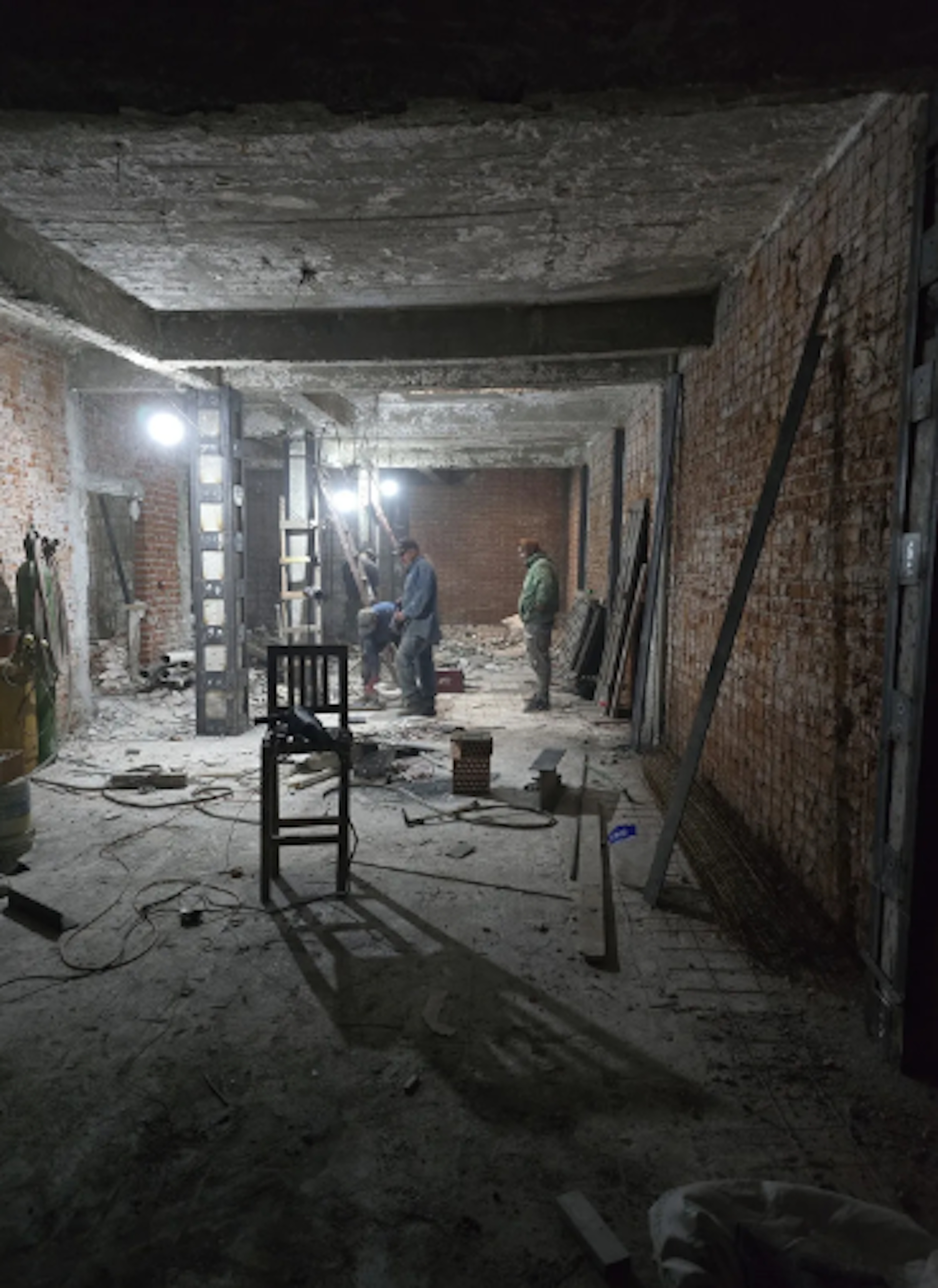 La imagen muestra un espacio interior en plena renovación. Se observan paredes de ladrillo expuesto, escombros en el suelo y trabajadores realizando labores. El techo está en mal estado y hay cables eléctricos visibles. Una silla solitaria se encuentra en primer plano. La iluminación es tenue, proporcionada por luces temporales. Es evidente que se está llevando a cabo una importante remodelación o restauración del inmueble.