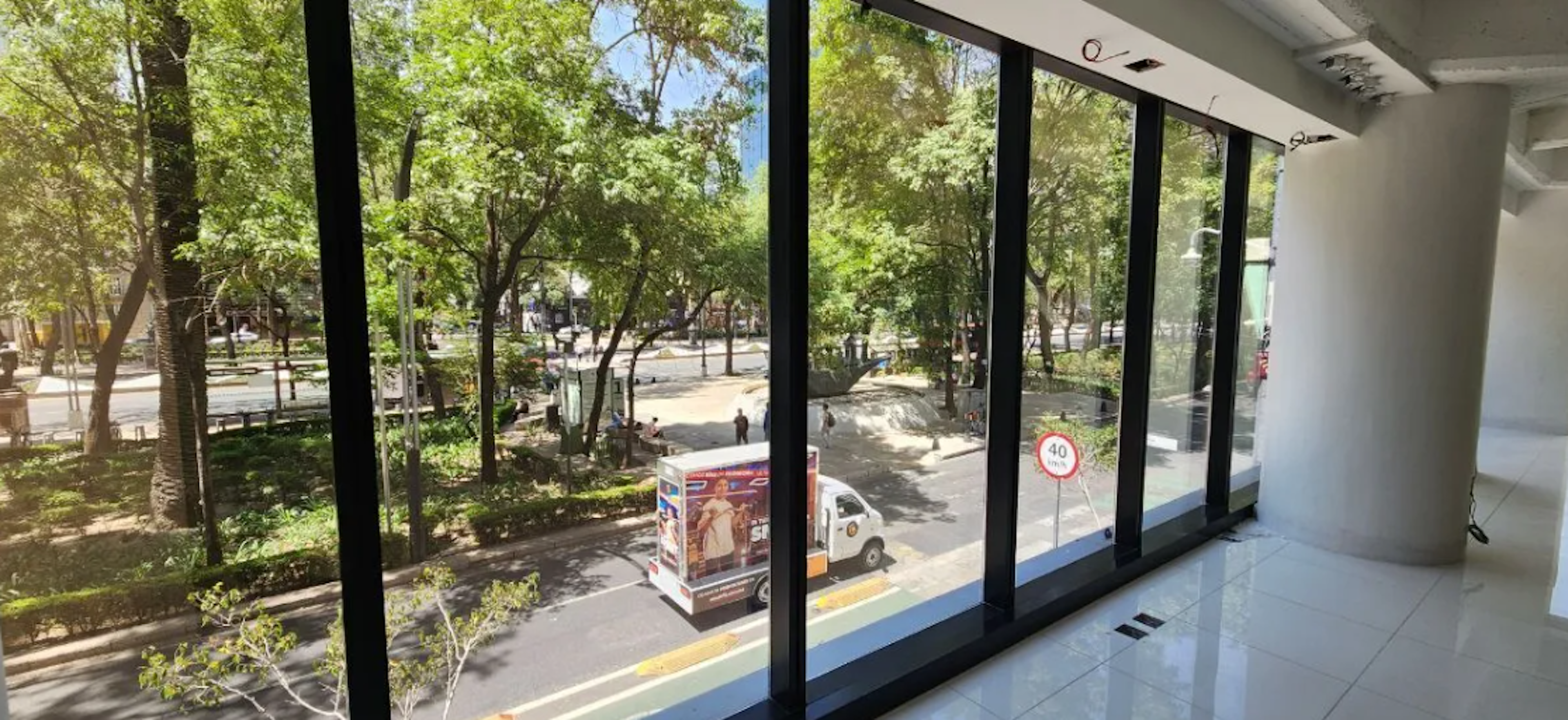 La imagen muestra una vista desde un interior moderno con grandes ventanales que dan a una calle arbolada y un parque. Se aprecia un ambiente urbano con áreas verdes, vehículos estacionados y señalización vial. La ubicación parece ser una zona céntrica y residencial con una mezcla de naturaleza y desarrollo urbano, ideal para propiedades con vistas agradables y acceso a espacios públicos.