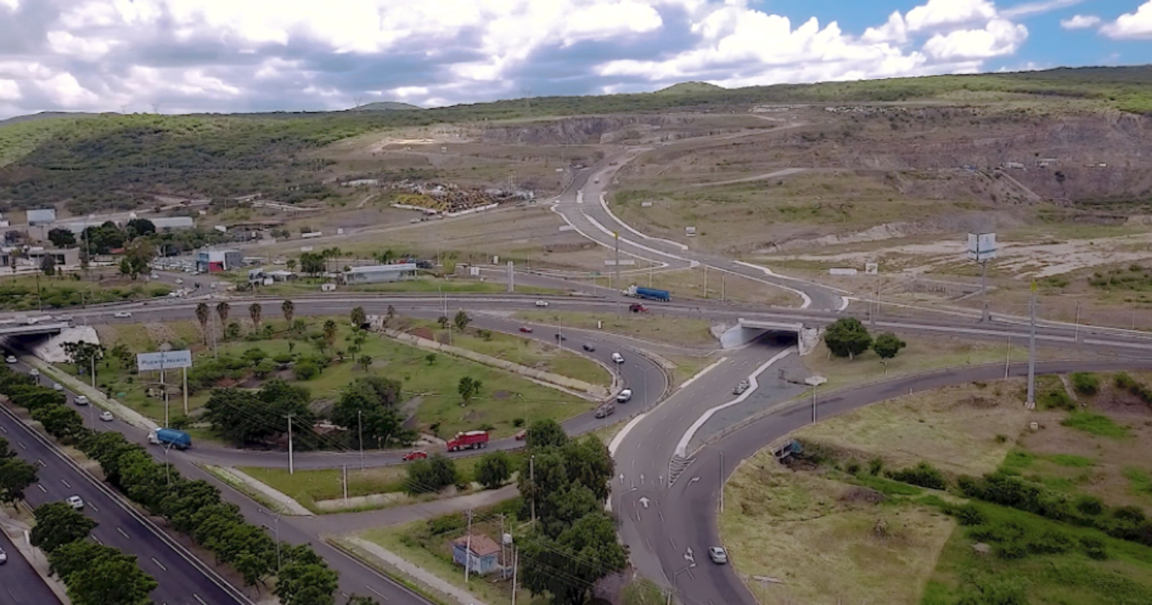 La imagen muestra una vista aérea de una intersección de carreteras en una zona semiurbana. Se observa un desarrollo de infraestructura vial con múltiples carriles, túneles y rotondas. El área circundante incluye algunas edificaciones, áreas verdes y colinas en el fondo. Este tipo de desarrollo podría indicar una zona en expansión con potencial para futuros proyectos inmobiliarios residenciales o comerciales.