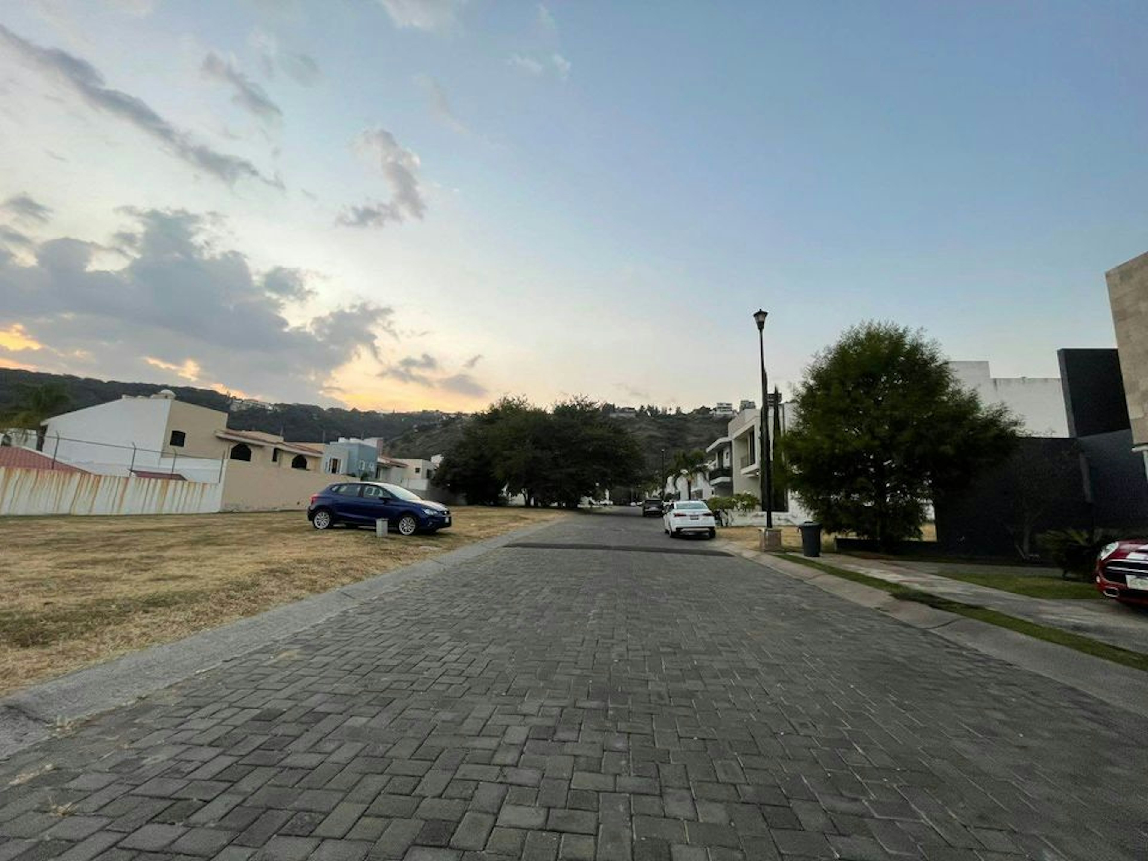 La imagen muestra una calle residencial tranquila con casas modernas y árboles. El pavimento es de adoquines y hay algunos autos estacionados. Se aprecia un cielo despejado con nubes suaves al atardecer. La zona parece ser un desarrollo inmobiliario reciente en una ubicación suburbana, con colinas visibles en el fondo. El ambiente general sugiere una comunidad residencial de clase media-alta.