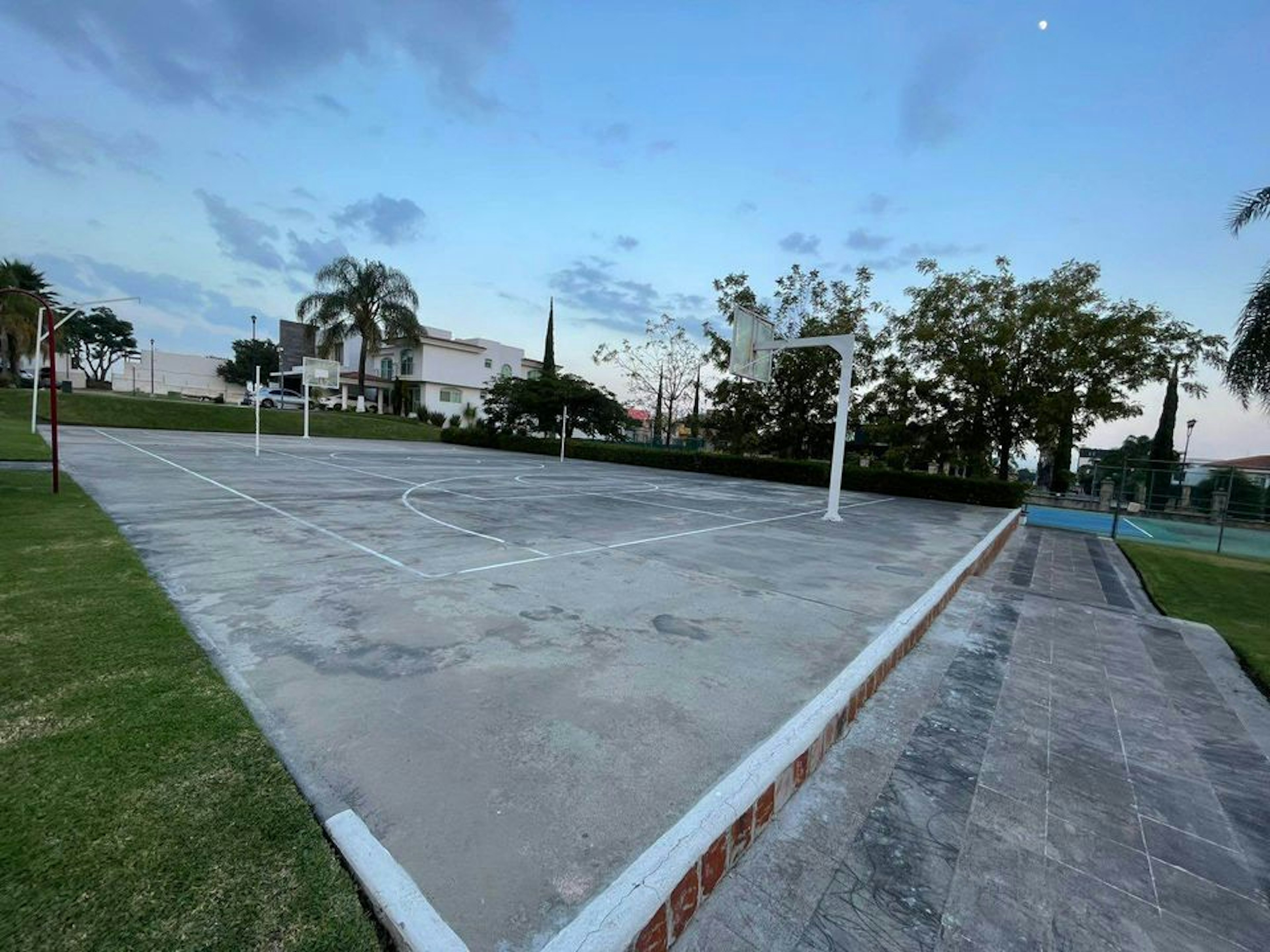 La imagen muestra una cancha de baloncesto al aire libre en un entorno residencial. La cancha está rodeada de césped y árboles, con casas blancas visibles en el fondo. El cielo es azul claro con algunas nubes. La cancha parece ser parte de una instalación comunitaria o un área recreativa en un vecindario de aspecto agradable, lo que sugiere que podría ser una amenidad valiosa para propiedades inmobiliarias cercanas.