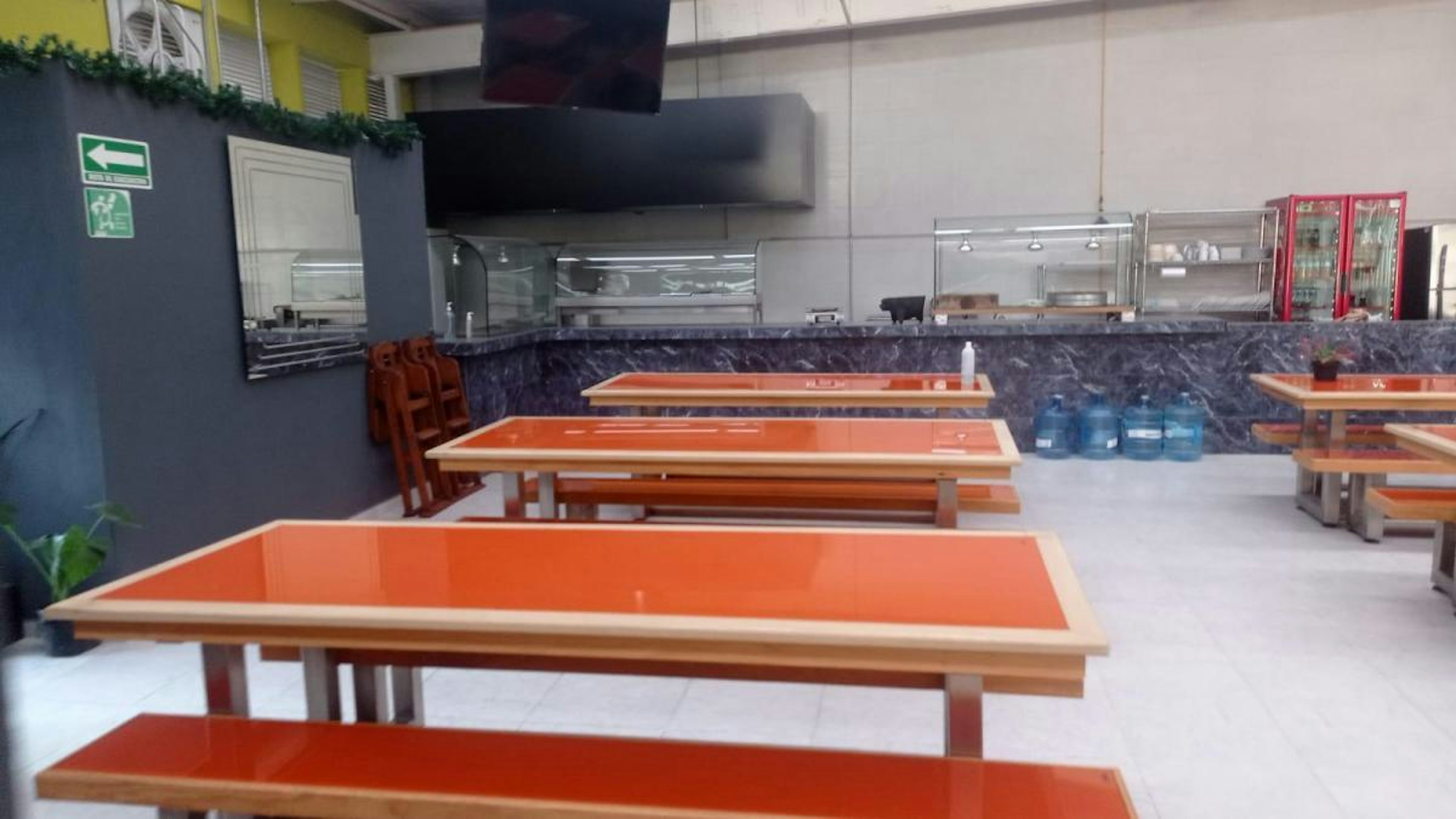 La imagen muestra un área de comedor o cafetería con mesas y bancos de color naranja. Al fondo se observa una barra de servicio con equipos de cocina y refrigeradores. Las paredes son de color gris oscuro con algunos detalles decorativos. Hay un televisor montado en la pared y señales de seguridad. El espacio parece ser parte de un establecimiento comercial o institucional, posiblemente una cafetería o comedor de empresa.