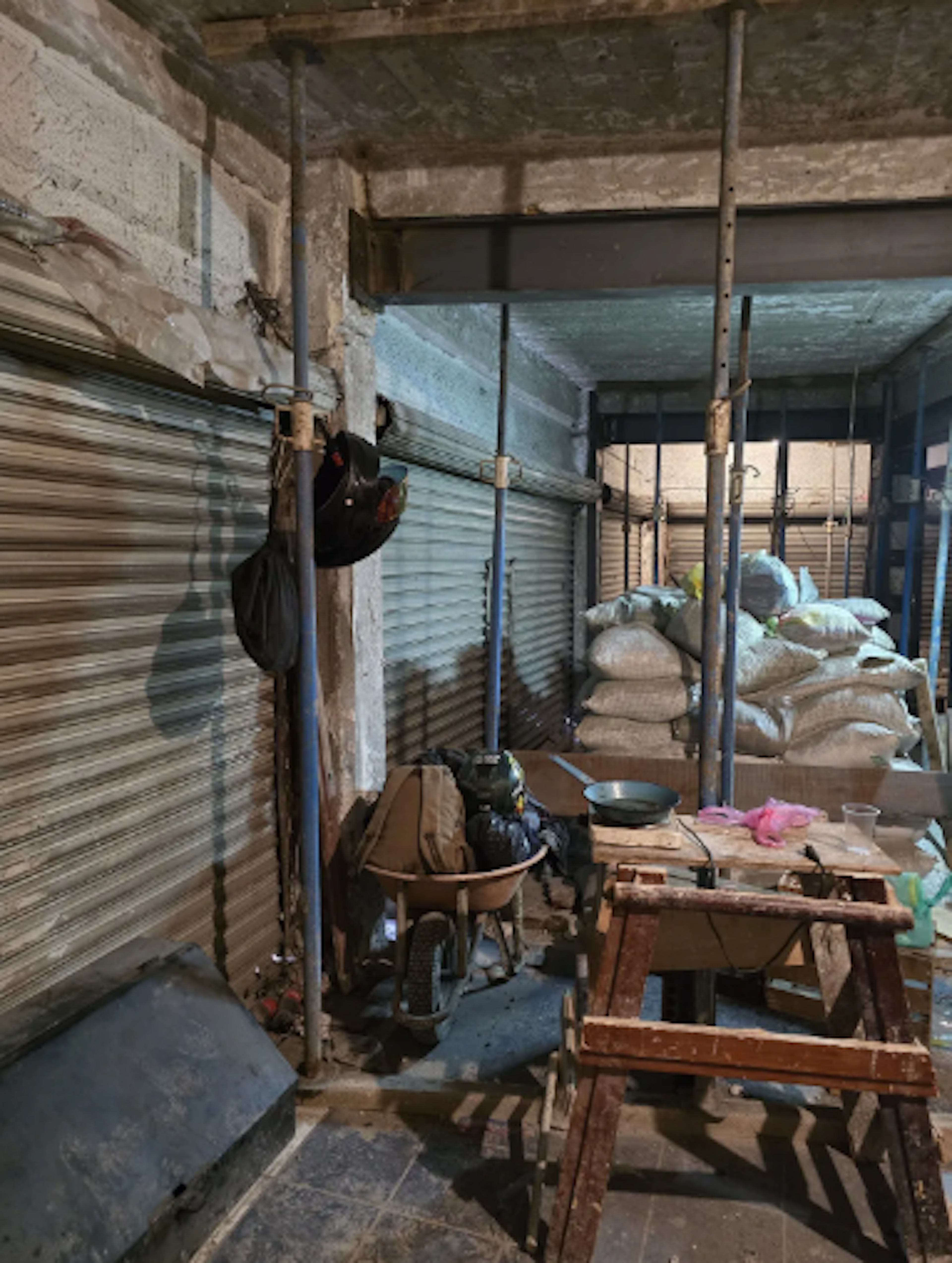 La imagen muestra un espacio de almacenamiento o área de trabajo en mal estado. Se observan paredes descuidadas, tuberías expuestas y una persiana metálica cerrada. Hay sacos apilados, una carretilla, herramientas y diversos objetos dispersos. El lugar parece abandonado o en proceso de renovación, indicando una propiedad que requiere rehabilitación sustancial para ser habitable o funcional.