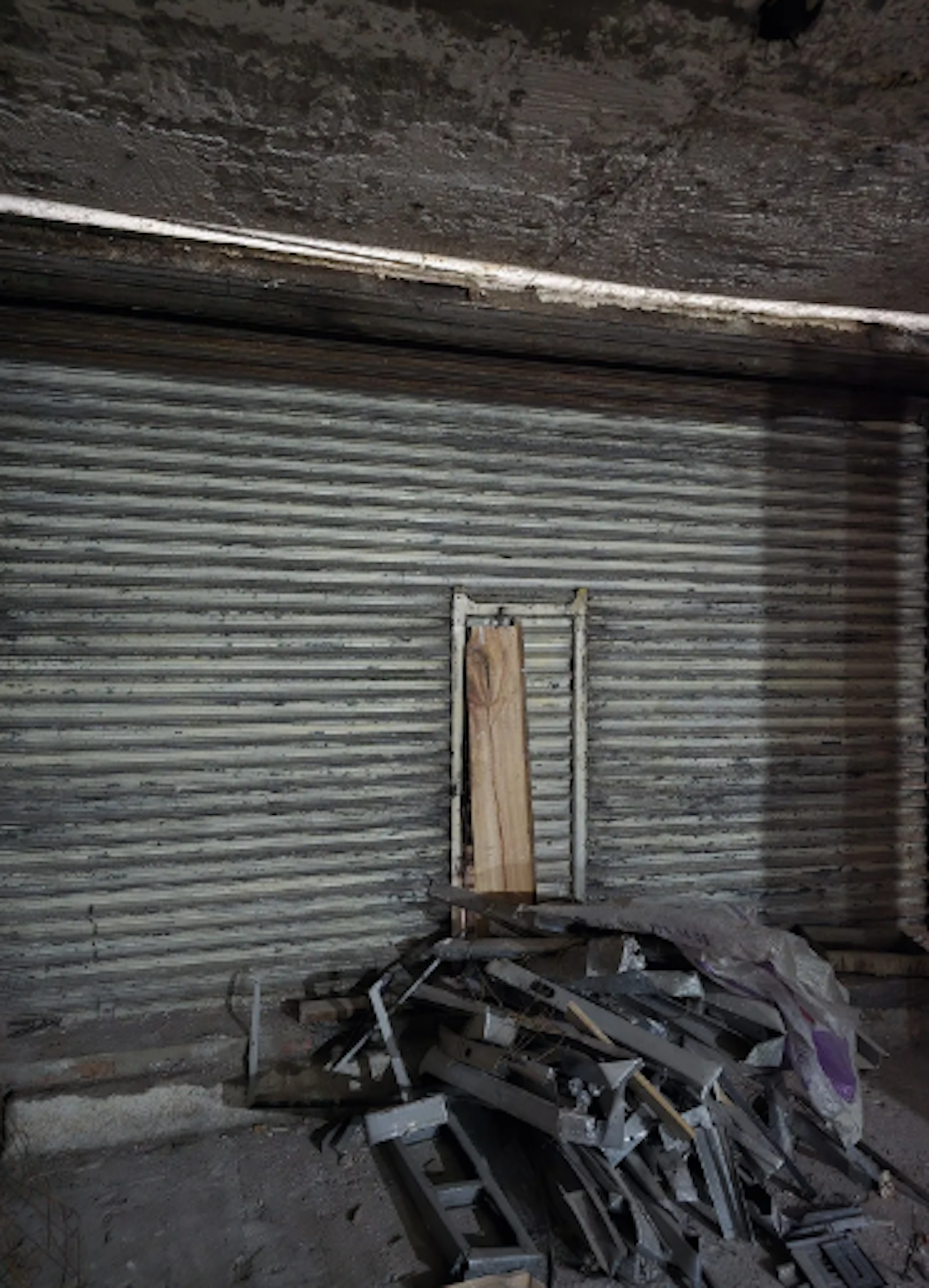 La imagen muestra un espacio interior deteriorado, posiblemente un garaje o almacén abandonado. Se observa una pared de metal corrugado con una pequeña abertura tapiada con madera. En el suelo hay un montón de escombros y materiales de construcción desechados. El techo parece estar en mal estado, con signos de desgaste y posible daño por agua. Esta escena sugiere un inmueble en estado de abandono o en necesidad de una renovación sustancial.