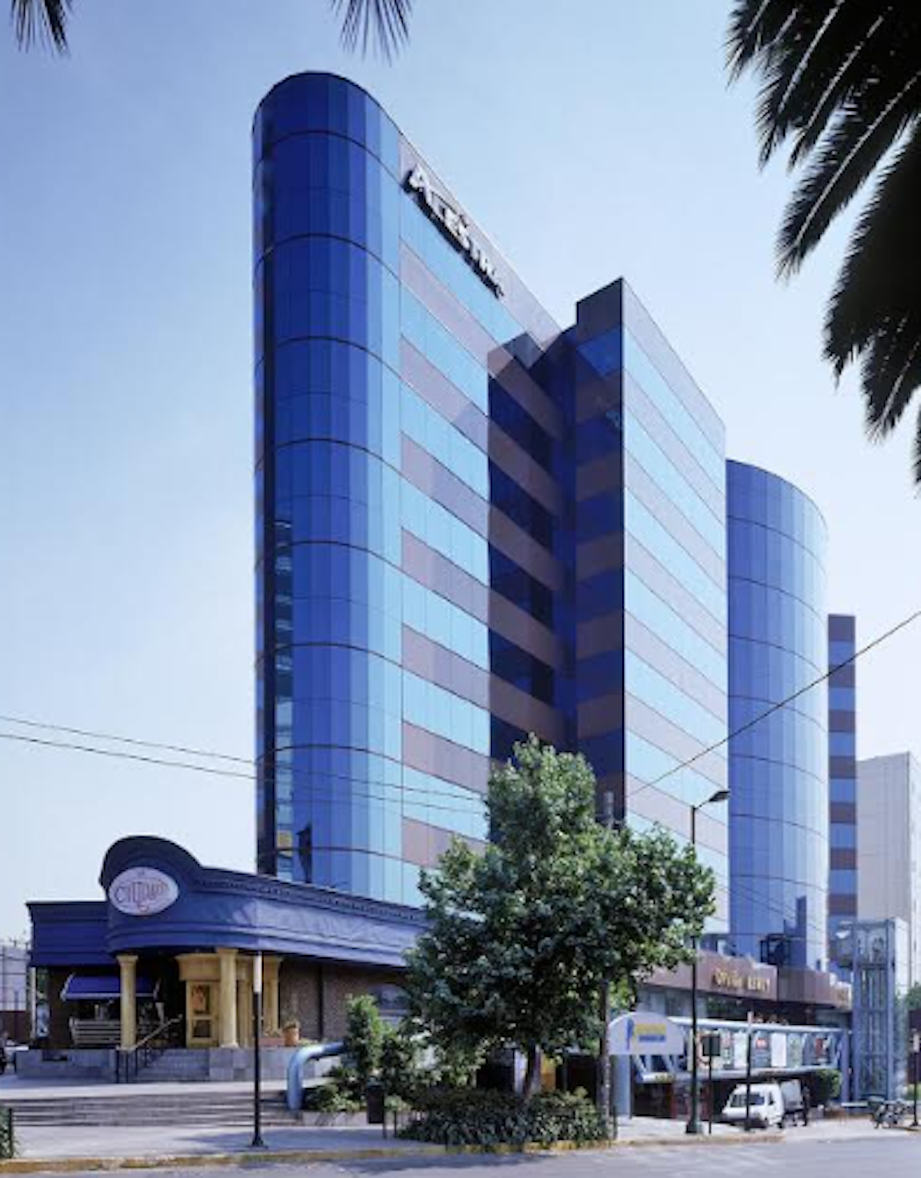 La imagen muestra un imponente edificio de oficinas moderno con fachada de cristal azul reflectante. El edificio tiene una forma distintiva con curvas y esquinas redondeadas. En la planta baja hay un local comercial con techo azul. La escena urbana incluye palmeras, árboles y mobiliario urbano, sugiriendo una ubicación en una zona comercial o de negocios en una ciudad con clima cálido.