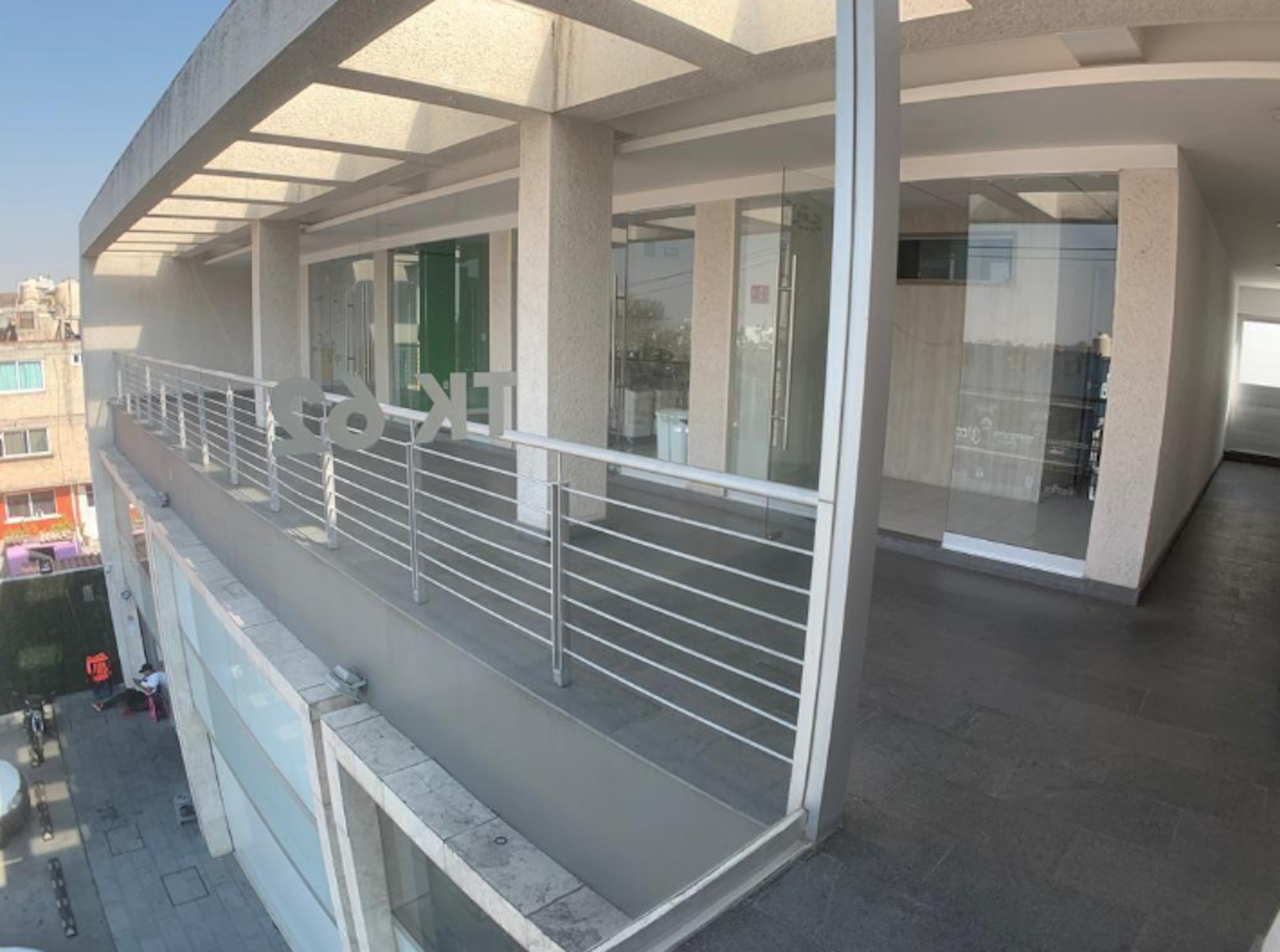 La imagen muestra la fachada de un edificio moderno, posiblemente un local comercial o de oficinas. Se aprecia un balcón o terraza en el segundo piso con barandillas metálicas. La estructura tiene grandes ventanales de vidrio, columnas blancas y un techo translúcido. El diseño es minimalista y funcional, típico de construcciones contemporáneas en el sector inmobiliario comercial.