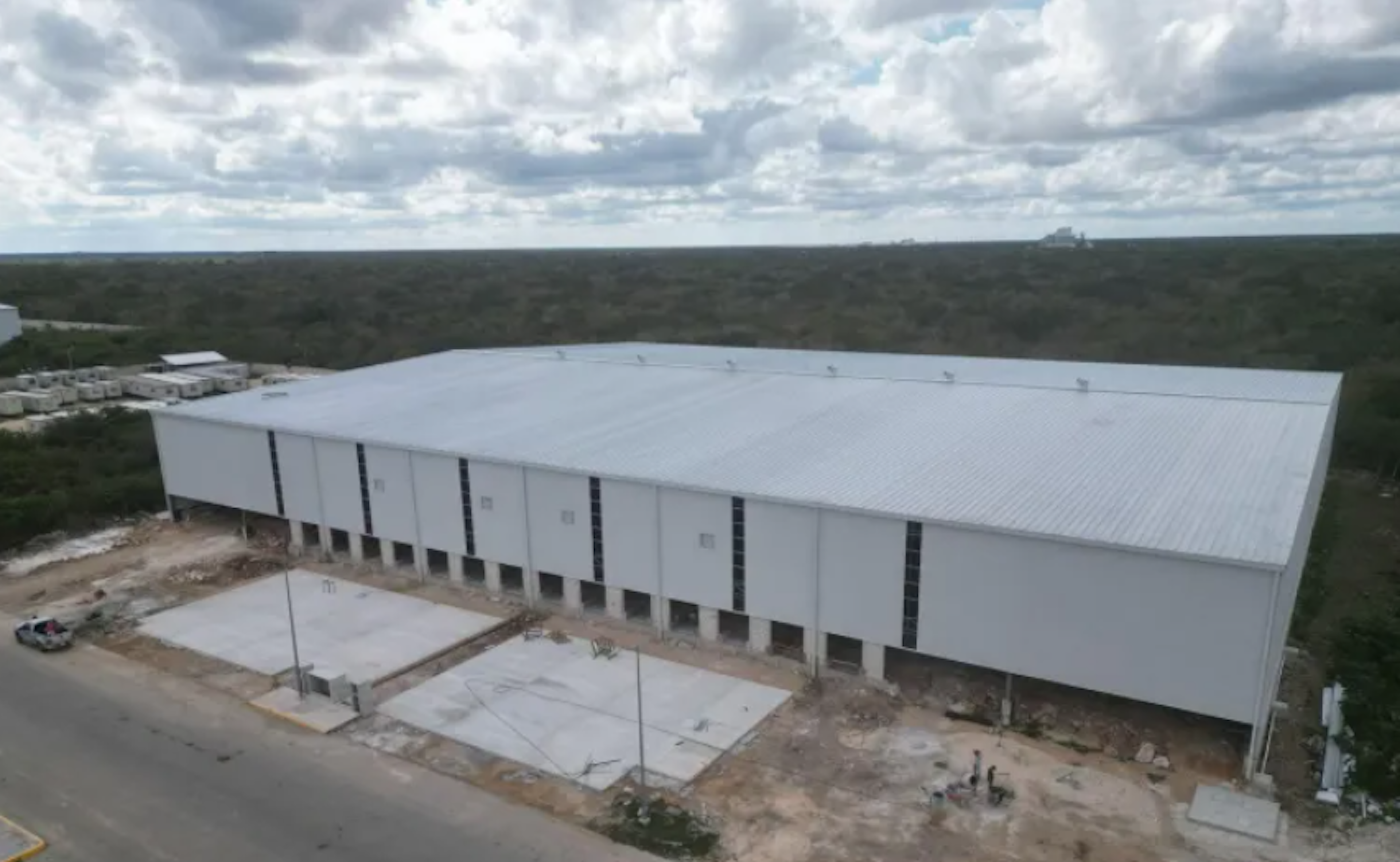 La imagen muestra una gran nave industrial o almacén de color blanco, ubicada en una zona rural o poco desarrollada. El edificio tiene múltiples puertas de carga, un techo amplio y plano, y está rodeado de un terreno sin pavimentar. Al fondo se observa un extenso bosque o vegetación densa. El cielo está nublado, dando un aspecto dramático a la escena. La estructura parece ser nueva o estar en construcción, dado el aspecto limpio y la presencia de áreas de trabajo alrededor.