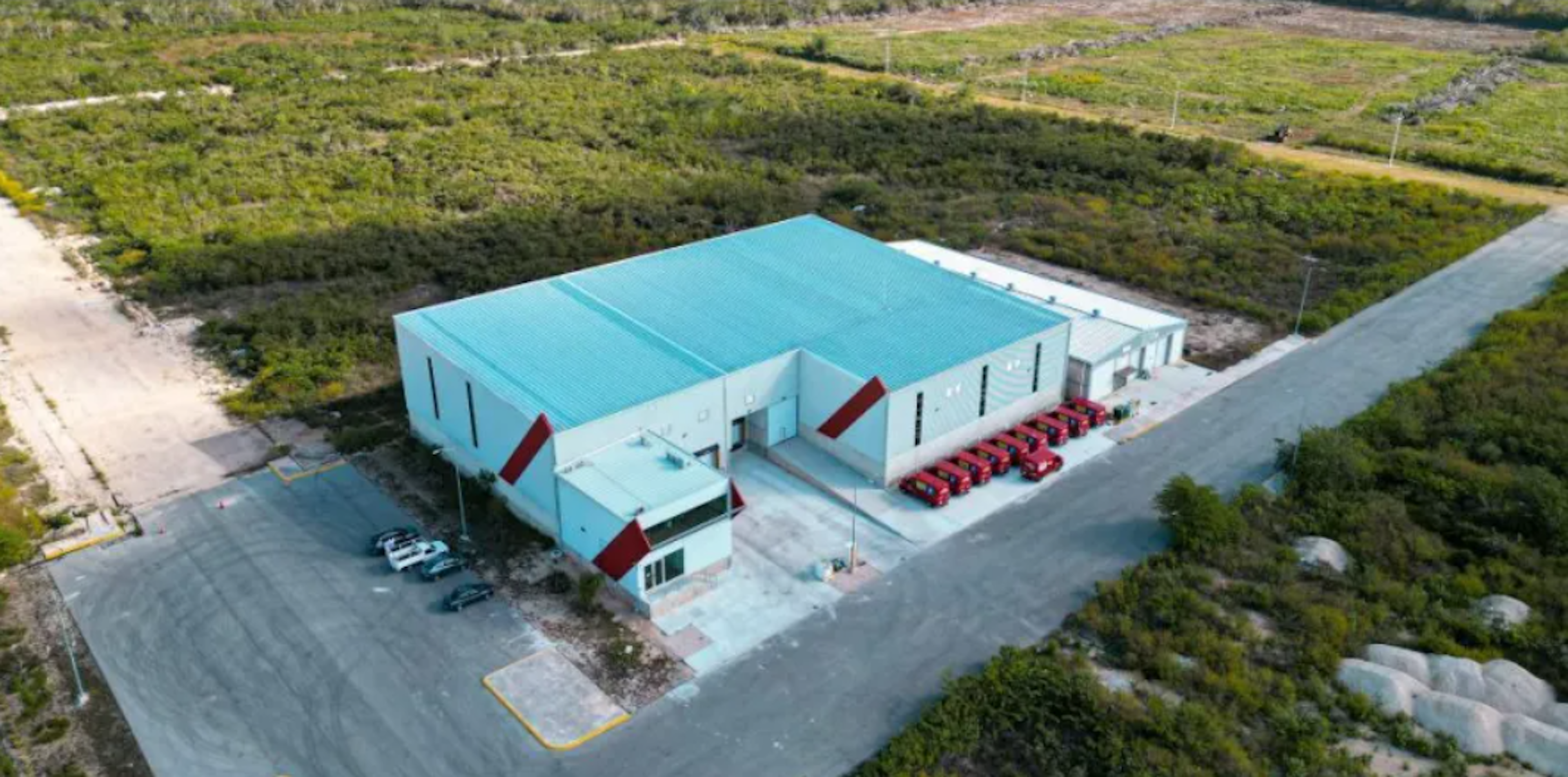 La imagen muestra una vista aérea de un edificio industrial de color turquesa con techo plano, ubicado en un área rural o suburbana. El edificio tiene forma rectangular y cuenta con un estacionamiento donde se observan varios vehículos rojos, posiblemente de la empresa. Está rodeado de vegetación y terrenos sin desarrollar, con caminos de acceso pavimentados. La estructura parece ser un almacén o centro de distribución moderno y bien mantenido.