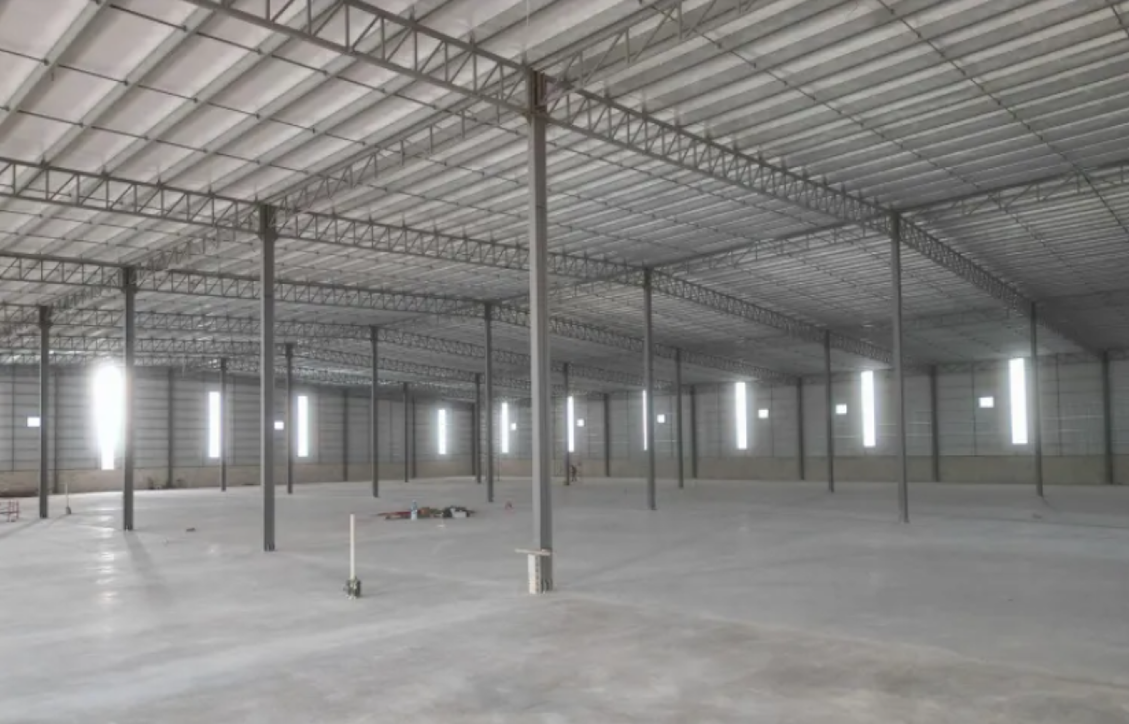 The image shows a vast, empty warehouse or industrial space under construction. It features a high ceiling supported by numerous metal beams and trusses, creating an expansive interior. The concrete floor is bare, and vertical support columns are evenly spaced throughout. Large, rectangular windows along the walls allow natural light to enter. The space appears to be in the final stages of construction, ready for customization or installation of equipment.
