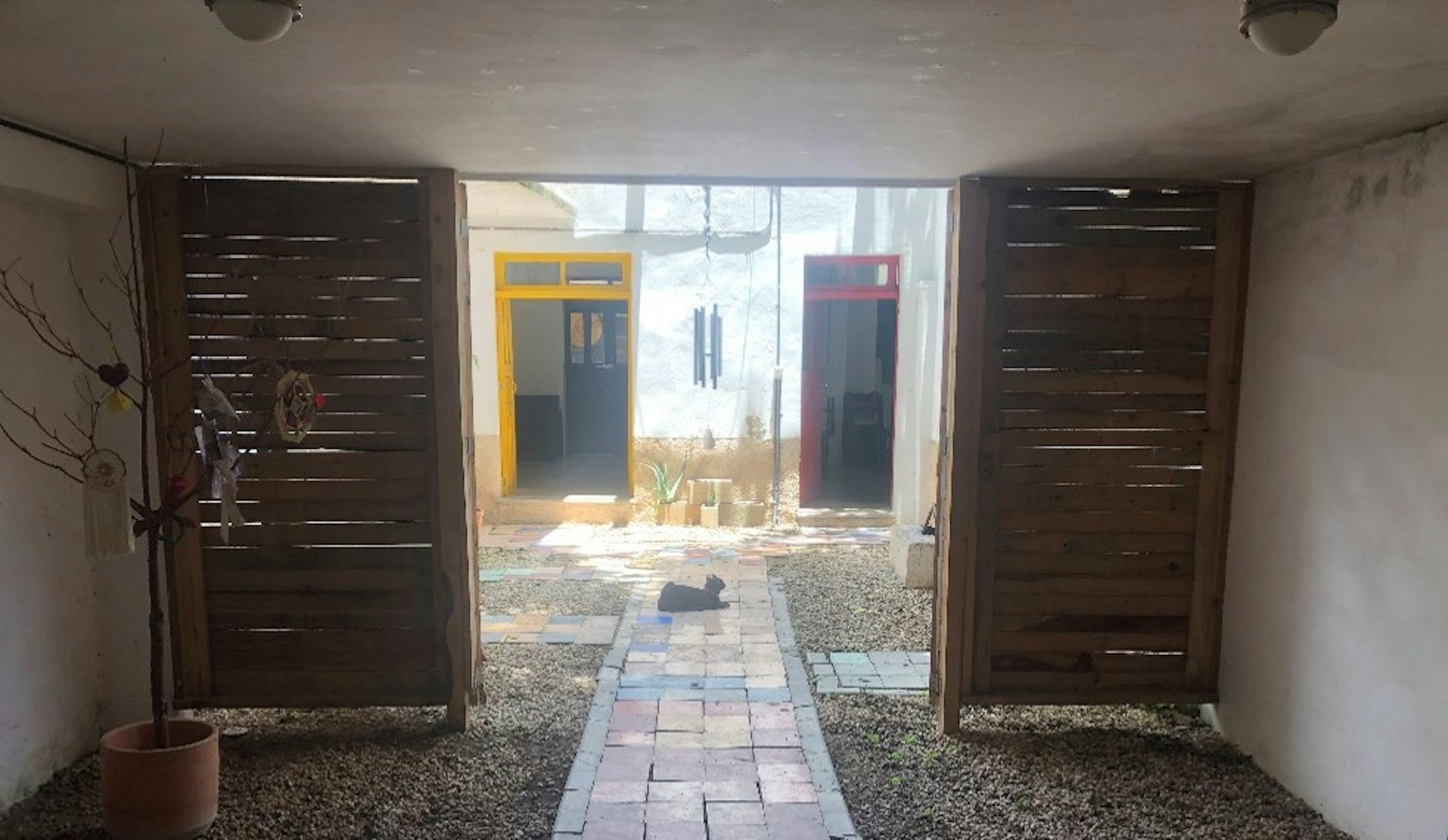 Esta imagen muestra un patio interior de una propiedad inmobiliaria, con puertas de madera abiertas que conducen a un espacio con suelo de adoquines y puertas coloridas al fondo. Un gato descansa en el camino.