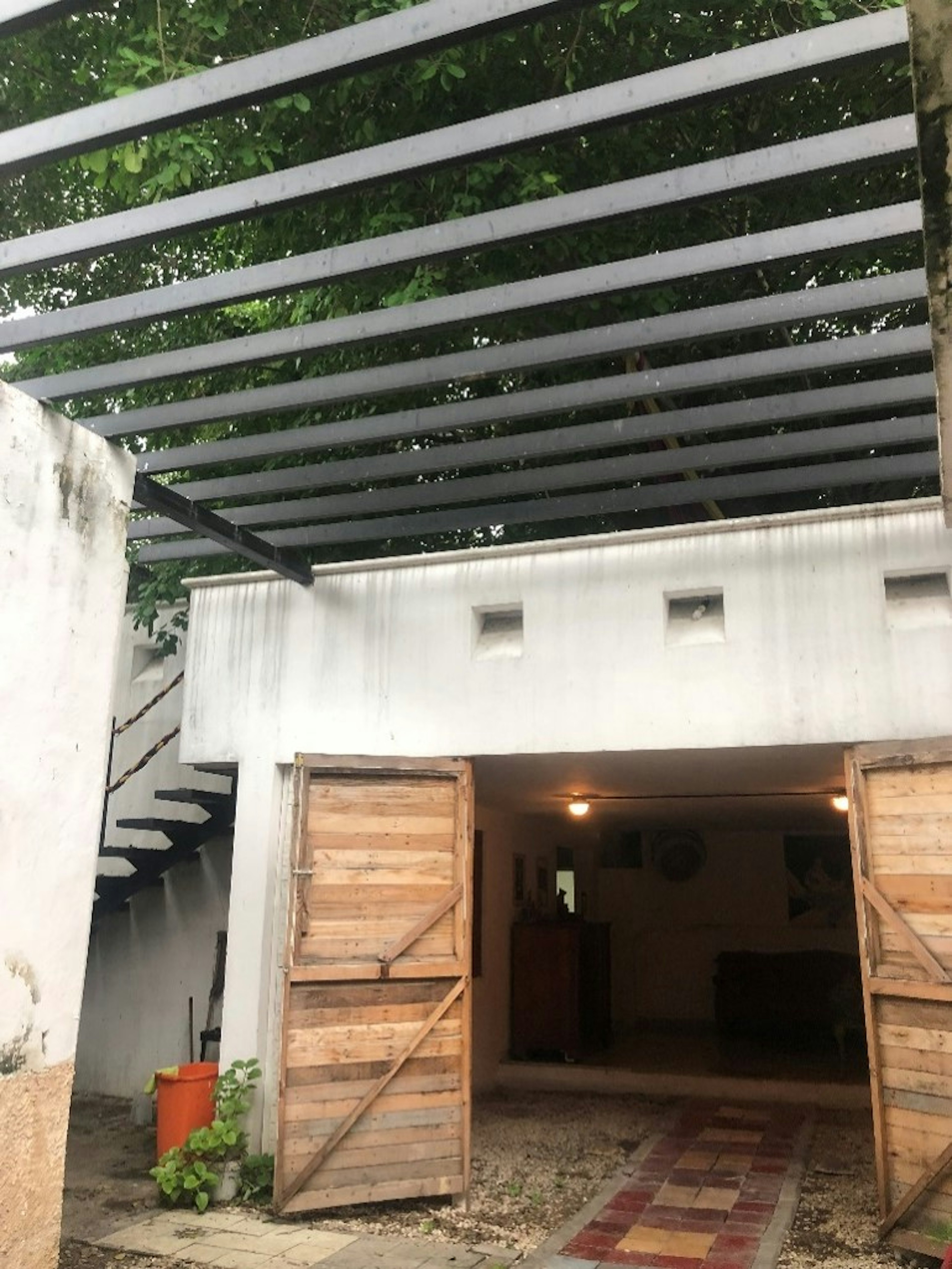 Esta imagen muestra la entrada de una propiedad con un diseño rústico. Se observa una pérgola de listones de madera, puertas de madera en estilo granero y una escalera al fondo que sugiere un segundo nivel. La vegetación añade un toque natural al entorno.