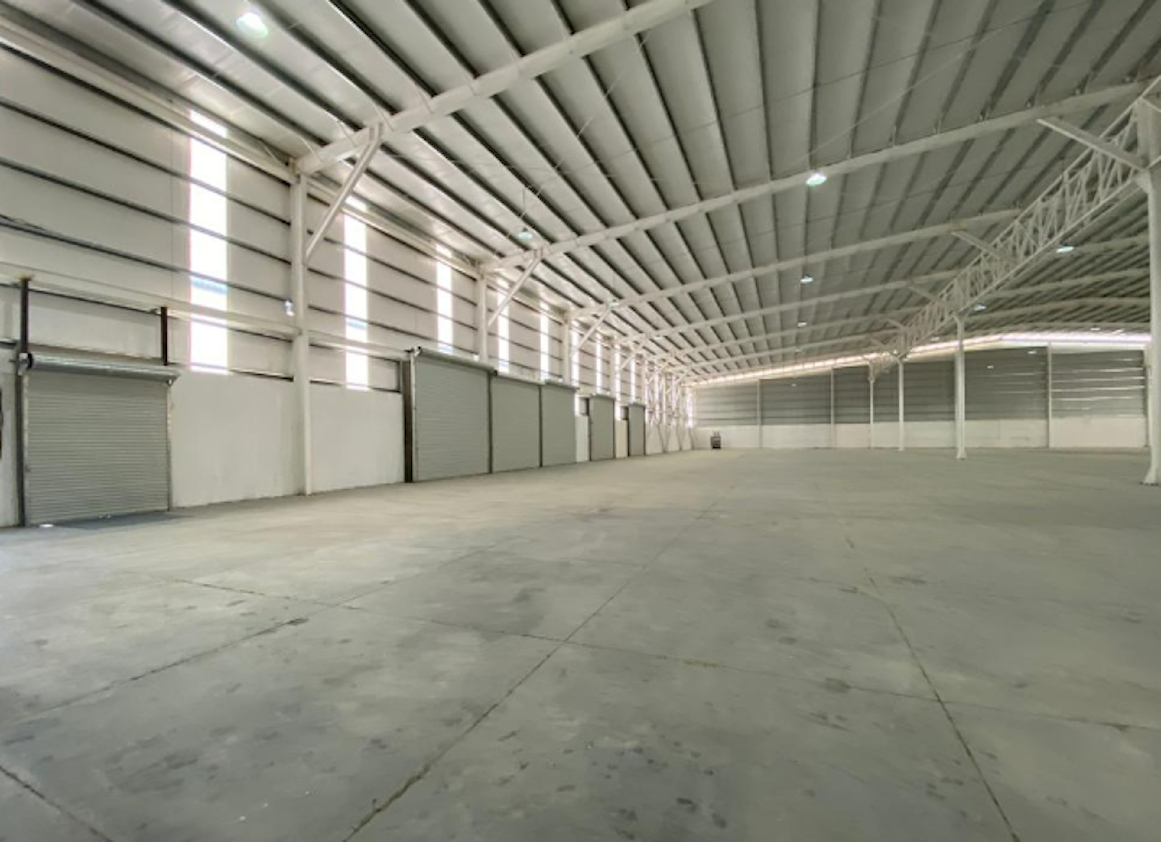 La imagen muestra el interior de una amplia bodega industrial vacía. El espacio cuenta con un techo alto de estructura metálica, paredes blancas y un piso de concreto. Se observan algunas puertas enrollables cerradas en un lateral. La iluminación natural ingresa por ventanas altas, complementada con iluminación artificial. El espacio diáfano ofrece gran capacidad de almacenamiento o potencial para diversos usos industriales o logísticos.