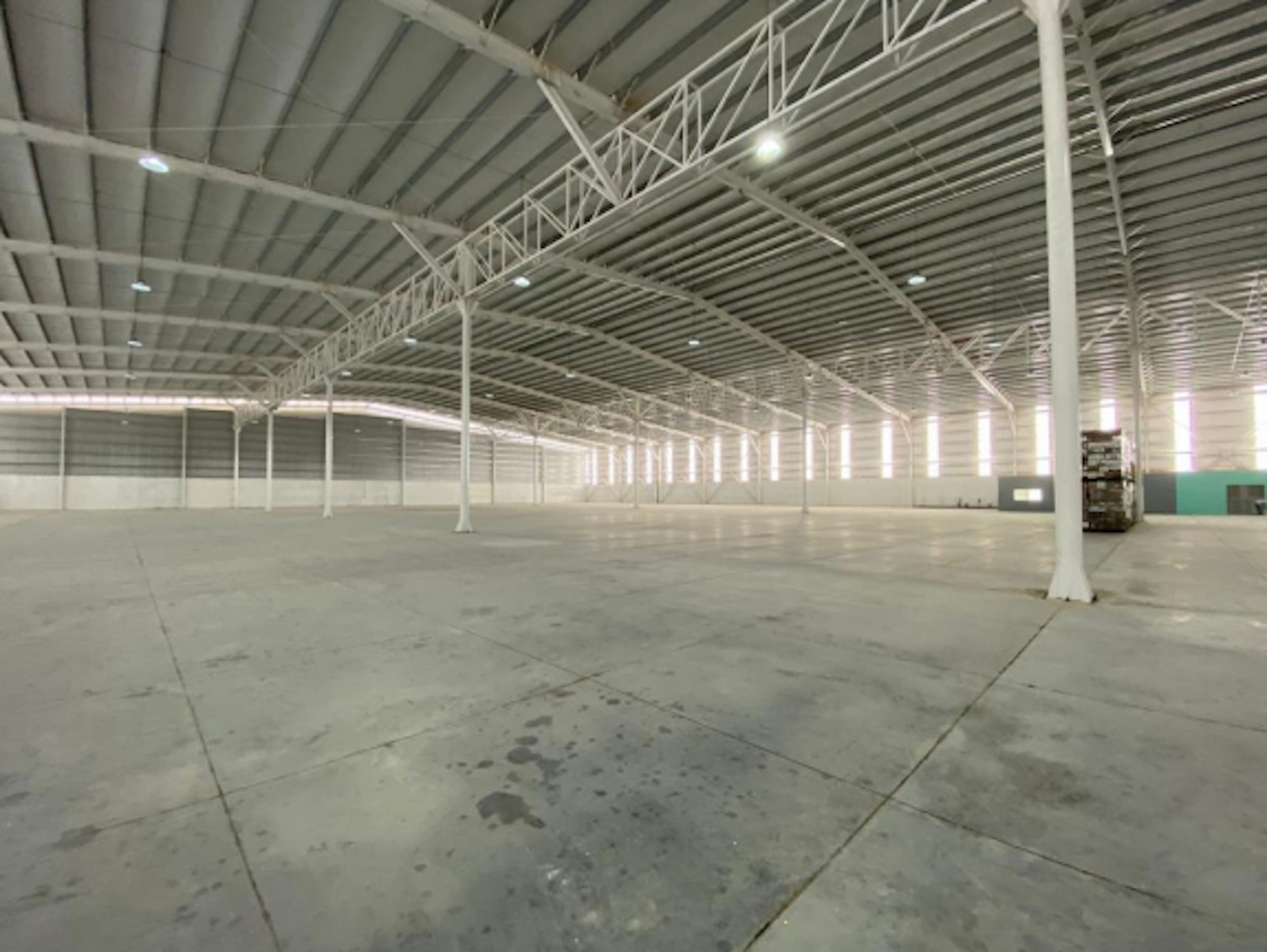 La imagen muestra un amplio almacén o nave industrial vacía con un techo alto de estructura metálica. El espacio es extenso, con suelo de concreto y columnas blancas que sostienen el techo. La iluminación proviene de luces LED instaladas en la estructura superior. Las paredes laterales son blancas y se observa una sección pintada de color turquesa al fondo. El espacio parece estar listo para ser utilizado o alquilado como área de almacenamiento o producción industrial.