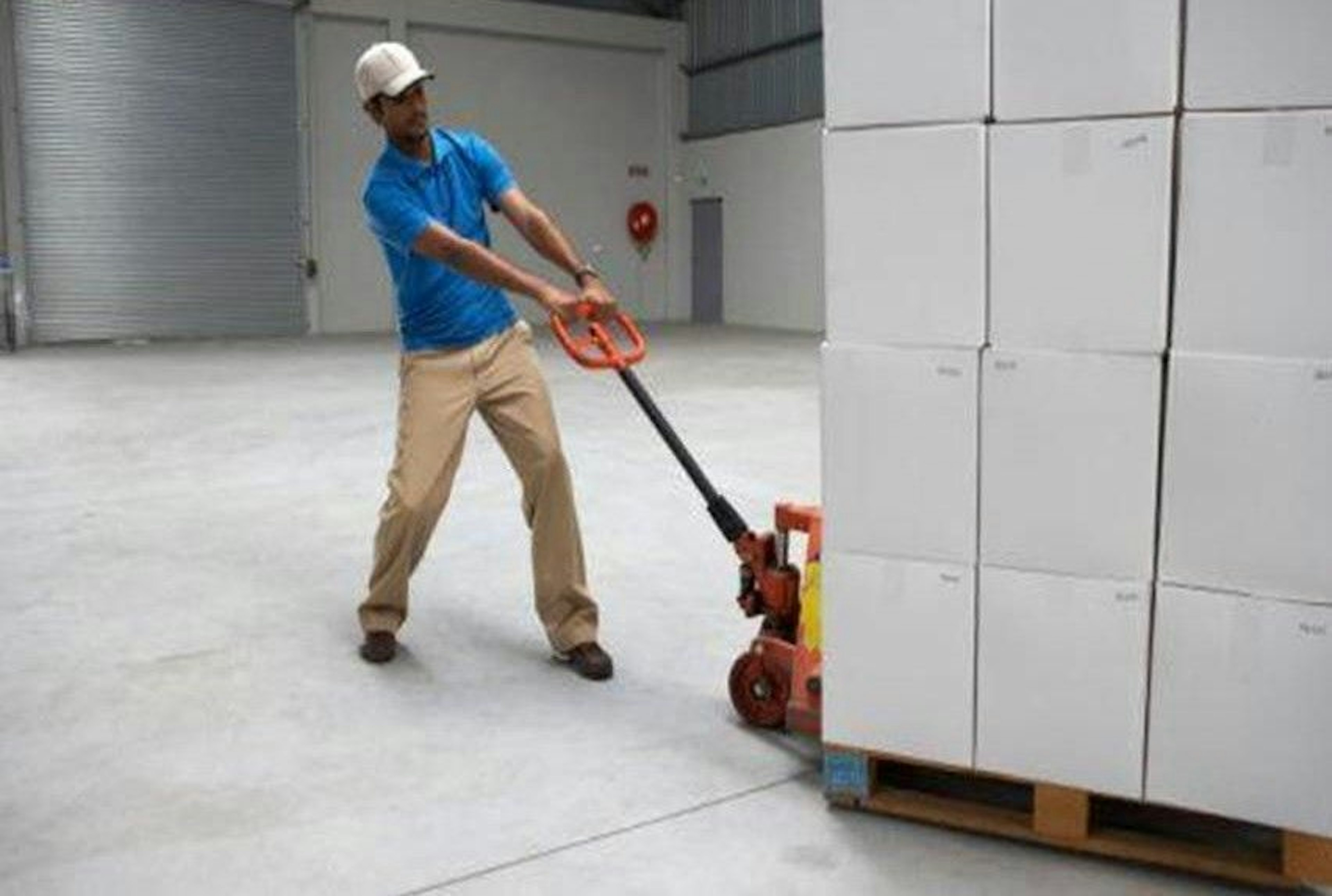 La imagen muestra a un trabajador en un almacén o centro de distribución utilizando un gato hidráulico manual para mover una pila de cajas blancas sobre un pallet. El empleado viste una camisa azul, pantalones beige y una gorra blanca. El espacio es amplio con paredes blancas y piso de concreto, típico de instalaciones logísticas o industriales.