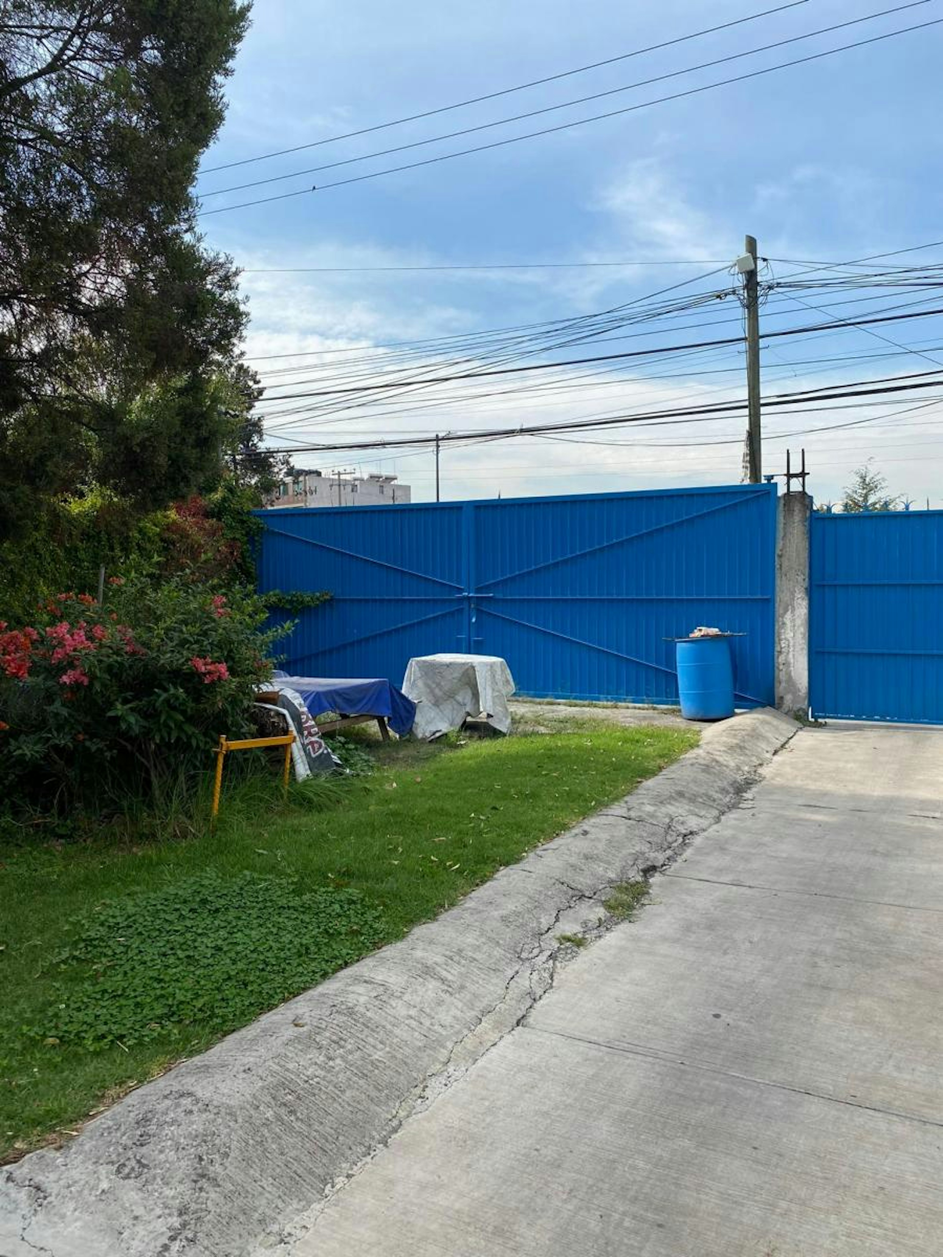 Imagen muestra un terreno con césped y vegetación, cercado por una valla metálica azul. Hay una silla plegable y objetos cubiertos con lonas.